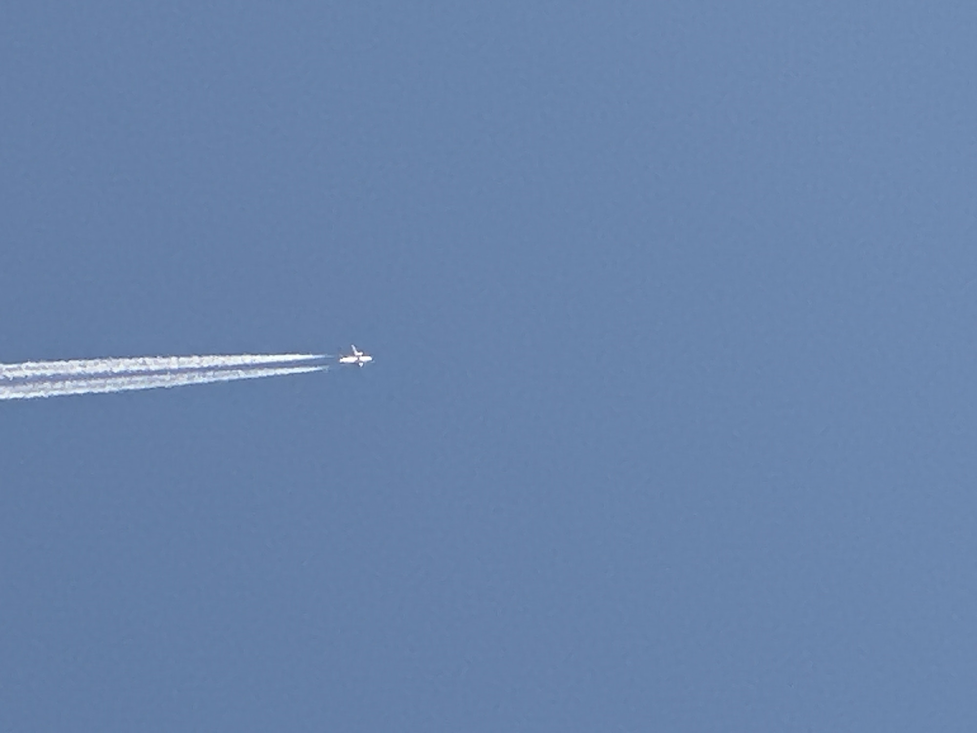 A small, white airplane is captured flying high across a clear, bright blue sky. The aircraft, positioned in the upper-left portion of the frame, is leaving a prominent, multi-stranded white contrail (condensation trail) that stretches horizontally behind it. The contrail extends from the plane's engines towards the left edge of the image, gradually dissipating into the sky.
The scene depicts an unclouded, expansive blue sky, indicative of daytime and fair weather conditions. The lighting is even and bright, suggesting a sunny environment. There are no other objects, geographical features, or clouds visible in the image, emphasizing the isolation of the plane against the vast sky.
The activity is simply an airplane in flight, producing a visible contrail at altitude. No people are discernible, and no specific emotions are conveyed. There is no visible text within the image. While the location context of Minsk, Belarus is provided, there are no visual cues within the image itself to confirm this geographical detail.