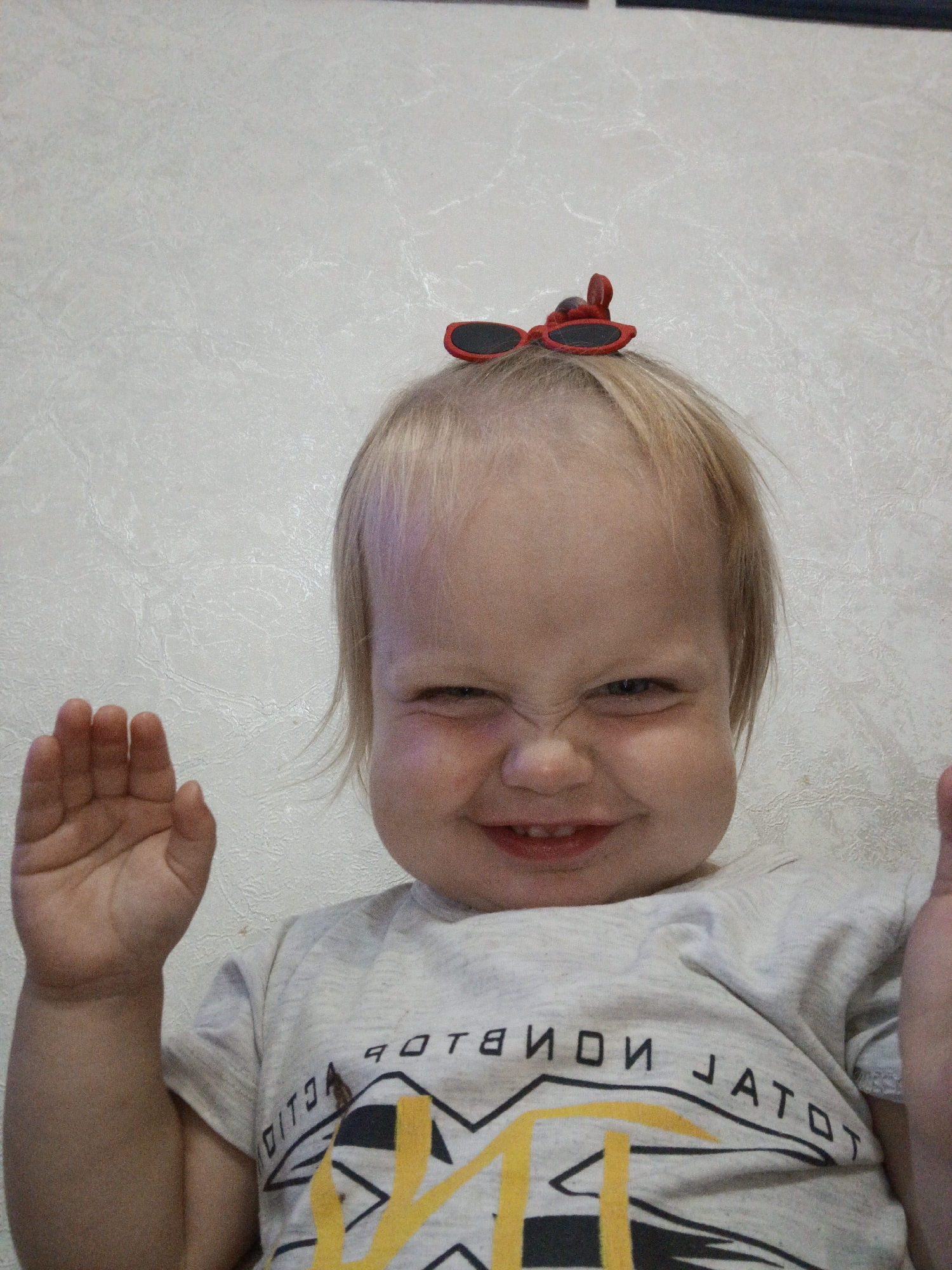 A young child with blonde hair is smiling and making a funny face while wearing red novelty sunglasses. The child is wearing a light-colored t-shirt with a graphic design that includes text. The background is a textured, light-colored wall. The child's left hand is raised, palm facing the camera, as if waving. The overall tone of the image is playful and lighthearted, suggesting a casual indoor setting. The text on the t-shirt appears to be in reverse and reads "TOTAL VISION" with some additional graphic elements. The location context provided is Medvedyovka, Russia.