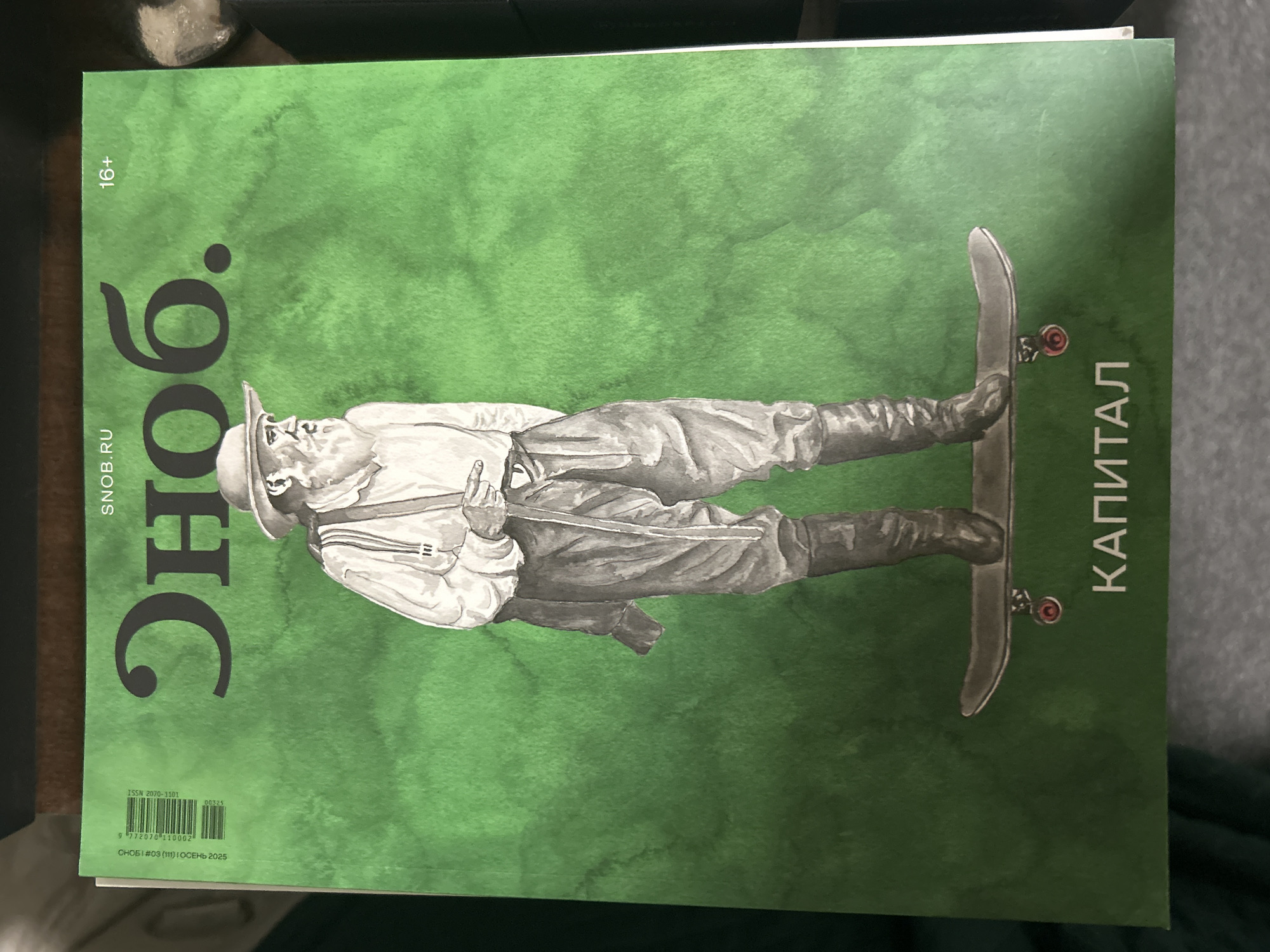 The image is a close-up, eye-level shot of the cover of a Russian magazine titled "СНОБ." (SNOb.). The magazine has a vibrant green, watercolor-like background. The cover features a black and white illustration of an elderly man with a long white beard and a hat, wearing suspenders, a shirt, and trousers, standing on a skateboard. The magazine's title, "СНОБ.", is prominently displayed in large, stylized black font at the top of the cover, to the right of a smaller "SNOB.RU". To the right of the title, "16+" is printed. In the bottom center of the cover, the word "КАПИТАЛ" (CAPITAL) is printed in white capital letters. The magazine's barcode and other publication information are visible on the left side of the cover. There is no discernible location in Moscow, Russia, as the image only shows the magazine cover.