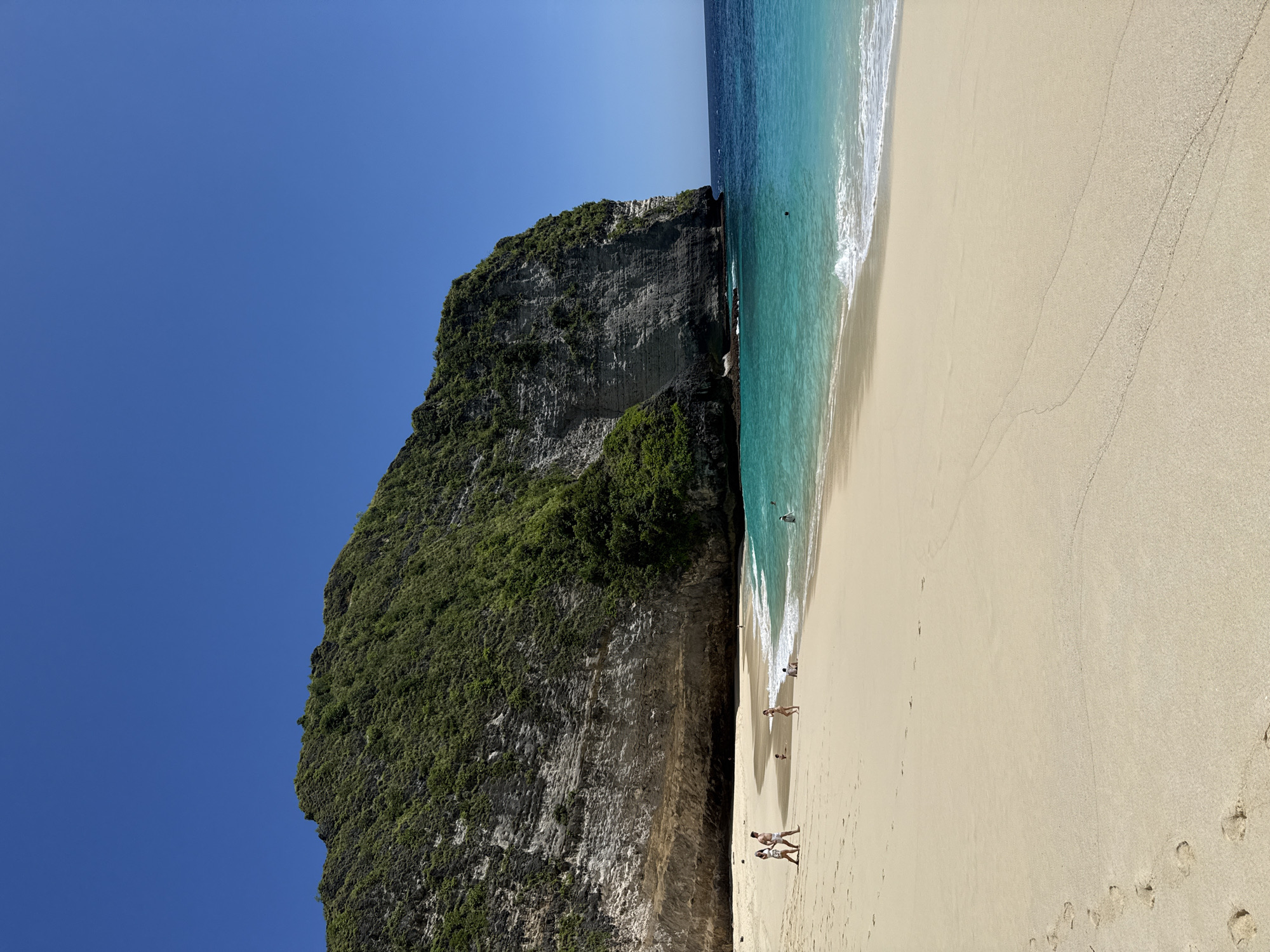 The image depicts a tropical beach scene in Bunga Mekar, Indonesia. A large, steep, rocky cliff covered with green vegetation rises sharply from the sand. To the right of the cliff, a sandy beach stretches along the turquoise ocean. Several people are walking along the shoreline, and one person appears to be swimming in the water. Footprints are visible in the sand. The sky is a clear, bright blue.