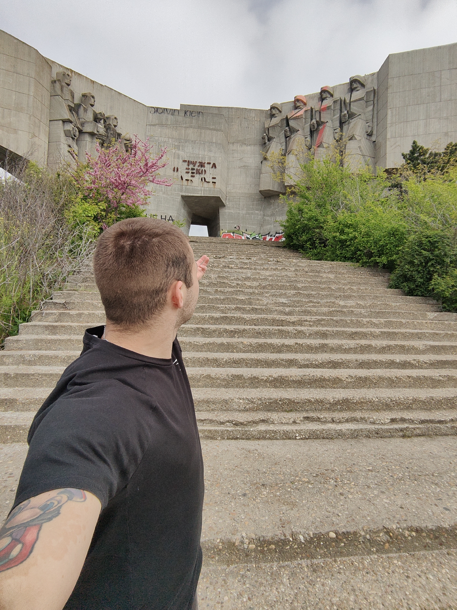 The image shows a man standing in front of the Monument to the Soviet Army in Varna, Bulgaria. The monument is a large concrete structure with carved figures of soldiers. The man is facing the monument and pointing towards it. The man has short brown hair and is wearing a black t-shirt. The staircase leading to the monument is in the foreground. The sky is overcast. There is graffiti on the monument, including the words "Calvin Klein". There is also some vegetation growing around the base of the monument.