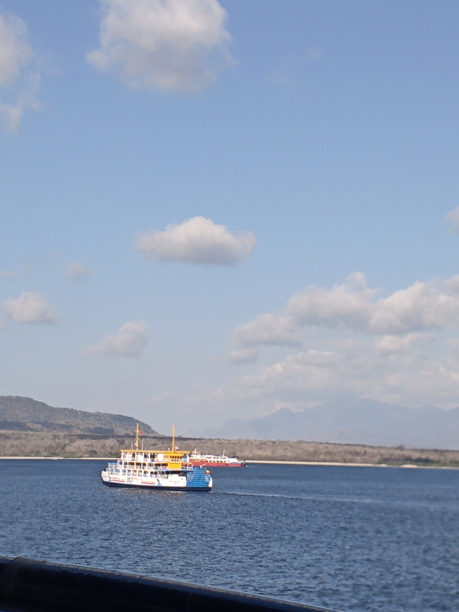A ferry boat with a white hull, blue lower deck, and yellow upper deck is moving through a body of water. The ferry has multiple decks and windows. Behind the ferry, another red and white ferry is visible in the distance, also on the water. The background shows a coastline with dry, scrubby vegetation and a hazy mountain range beyond. The sky is a light blue with scattered white clouds. The image appears to be taken from another vessel, with a dark railing visible in the foreground. The scene is consistent with a ferry crossing in a coastal area of Indonesia, possibly related to transportation between islands or across a strait.