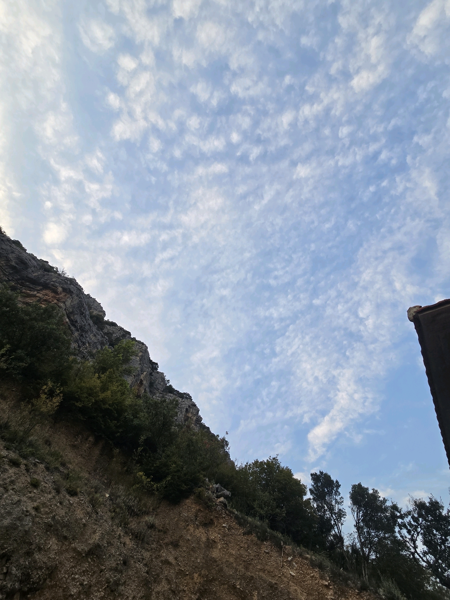 The image captures a rocky mountainside with sparse vegetation under a sky filled with wispy, scattered clouds. The foreground features dry, brown earth and rocks, with patches of green shrubs and trees growing along the slope. The mountain itself is steep and rugged, showing layers of rock and exposed soil. In the upper right corner, a portion of a dark-tiled roof and a brown structural edge are visible, suggesting the presence of a building near the mountainside. The lighting indicates it is daytime, likely late afternoon or early morning, given the soft light and the cloud formations. The scene is devoid of any human presence or activity. The location is not identifiable as being within the city of Podgorica, Montenegro.