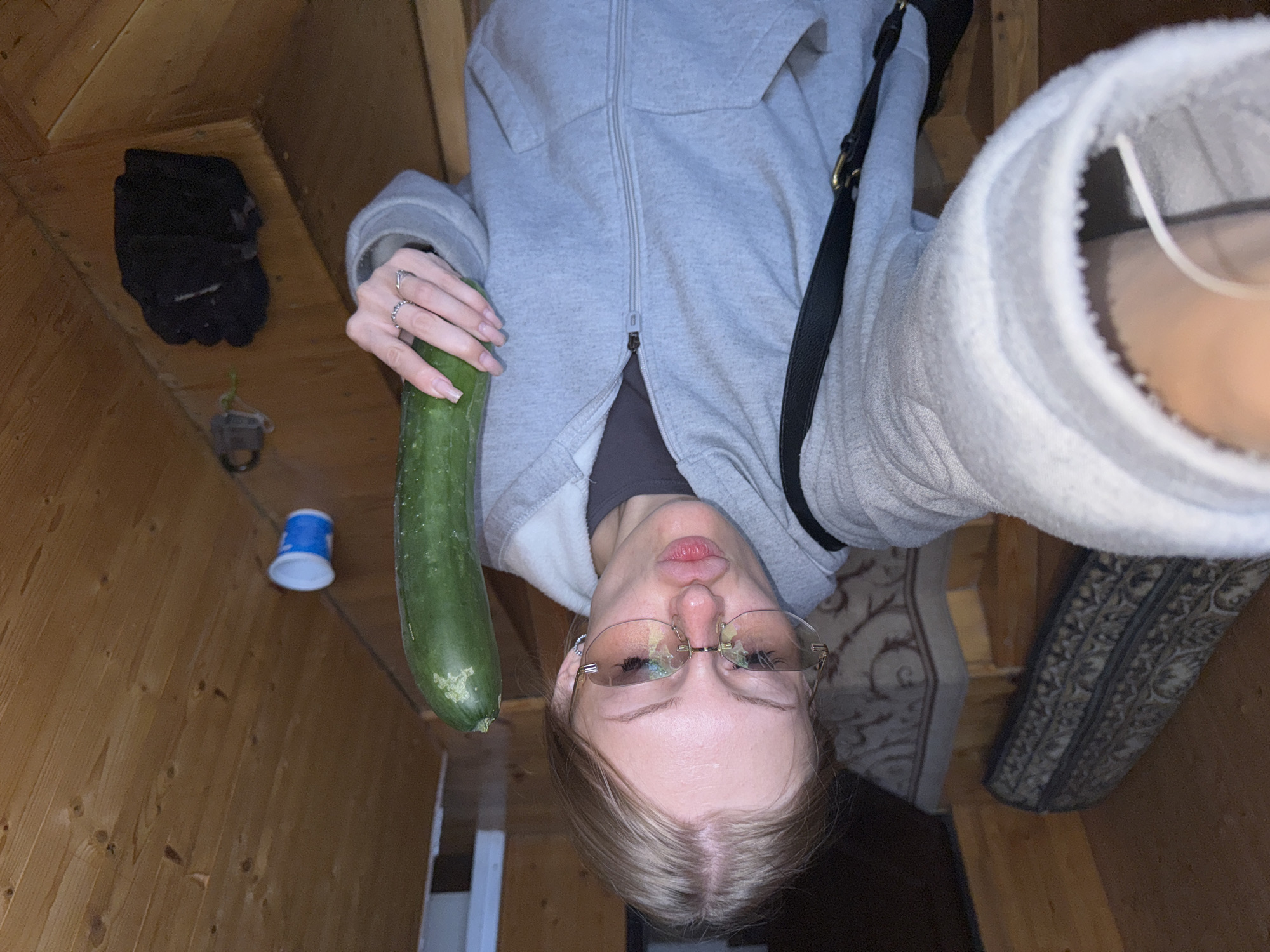 An overhead, slightly angled shot shows a young Caucasian woman indoors, possibly in an attic or loft space, given the exposed wooden beams of the ceiling and sloped walls. She is holding a long, green zucchini horizontally above her head with her right hand, her fingers adorned with rings. The woman is wearing a grey zip-up hoodie over a dark top and thin-rimmed glasses. Her face is turned towards the camera, with her lips slightly parted. In the background, on the wooden wall, a blue and white disposable cup is visible, along with a dark pair of gloves and a small grey object. The floor appears to be made of wood, with patterned rugs visible in the lower portion of the image. The overall lighting is dim, suggesting an indoor environment with limited light sources.