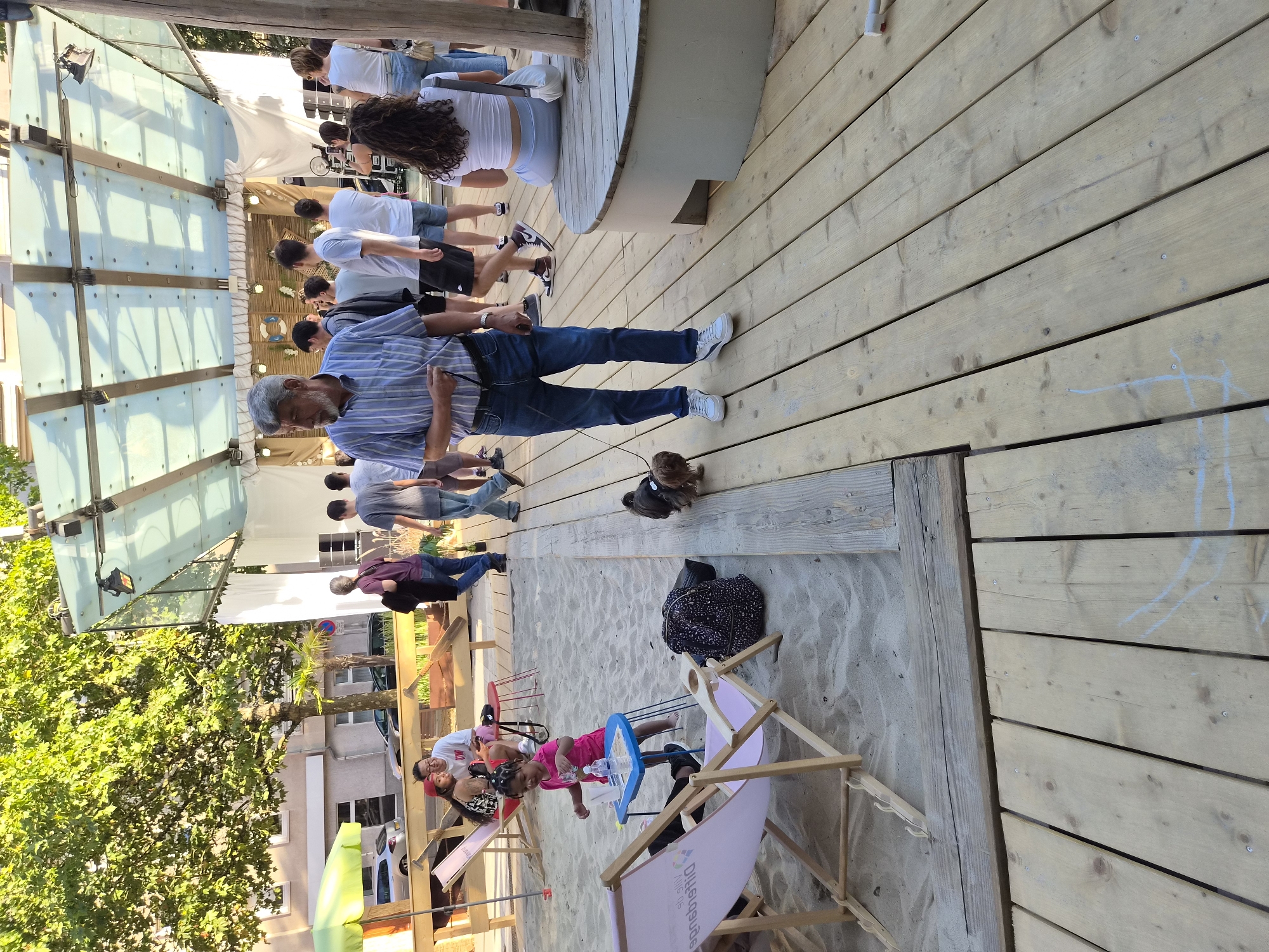 The image shows a scene in the city of Differdange, Luxembourg, at an outdoor event or venue. In the foreground, an older man with grey hair and a striped shirt is walking a small brown dog on a leash on a wooden deck. To the left, a sandy area features two pink lounge chairs, one occupied by a young child in a pink shirt reaching for a small blue table with a bottle of water and a cup. Further back, a young woman in a red cap and a man in a white shirt are sitting on a bench. In the background, a stage area is set up with a white backdrop, lights, and decorative elements, suggesting a performance or gathering is taking place. Several other people are visible in the midground and background, some standing and observing, others walking. The overall environment appears to be a public space designed for leisure and entertainment, with elements of sand, wood, and greenery from trees.