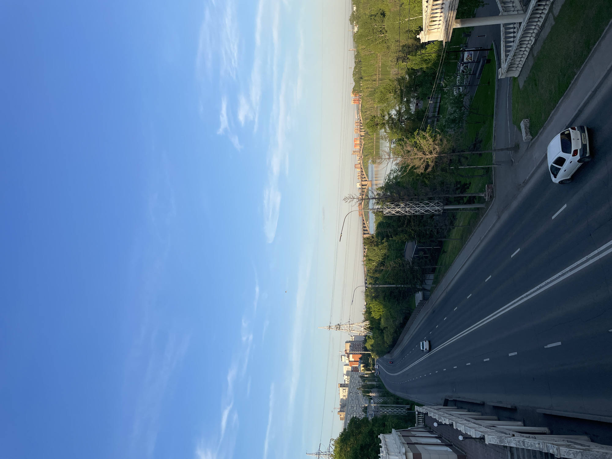 The image provides an elevated view of an urban landscape, likely from a bridge or overpass, under a clear, bright blue sky dotted with scattered, wispy white clouds. The setting sun casts a soft light across the scene, indicating late afternoon or early evening.
A wide, multi-lane asphalt road with prominent white lane markings curves into the distance, occupying the right side of the frame. A white car is parked on the shoulder of the road in the foreground, with its license plate partially visible, displaying "P738XO 124," with "124" being the regional code for Krasnoyarsk Krai, Russia. Further down the road, another white car is in motion, and distant red taillights suggest additional traffic. The elevated vantage point features a decorative white balustrade or railing along its edge.
To the left and across the midground, a significant body of water, presumably a river, flows, spanned by a multi-arched bridge. Lush green trees and vegetation line the riverbanks and parts of the road, suggesting a warm season. Utility poles and overhead power lines are visible throughout the composition, particularly on the left side of the road. In the background, across the river, several buildings are interspersed among the trees.
The scene depicts a calm, typical urban environment with light vehicular activity. No people are distinctly visible, and the overall mood is serene and uneventful. The clear weather and natural light contribute to a peaceful atmosphere.