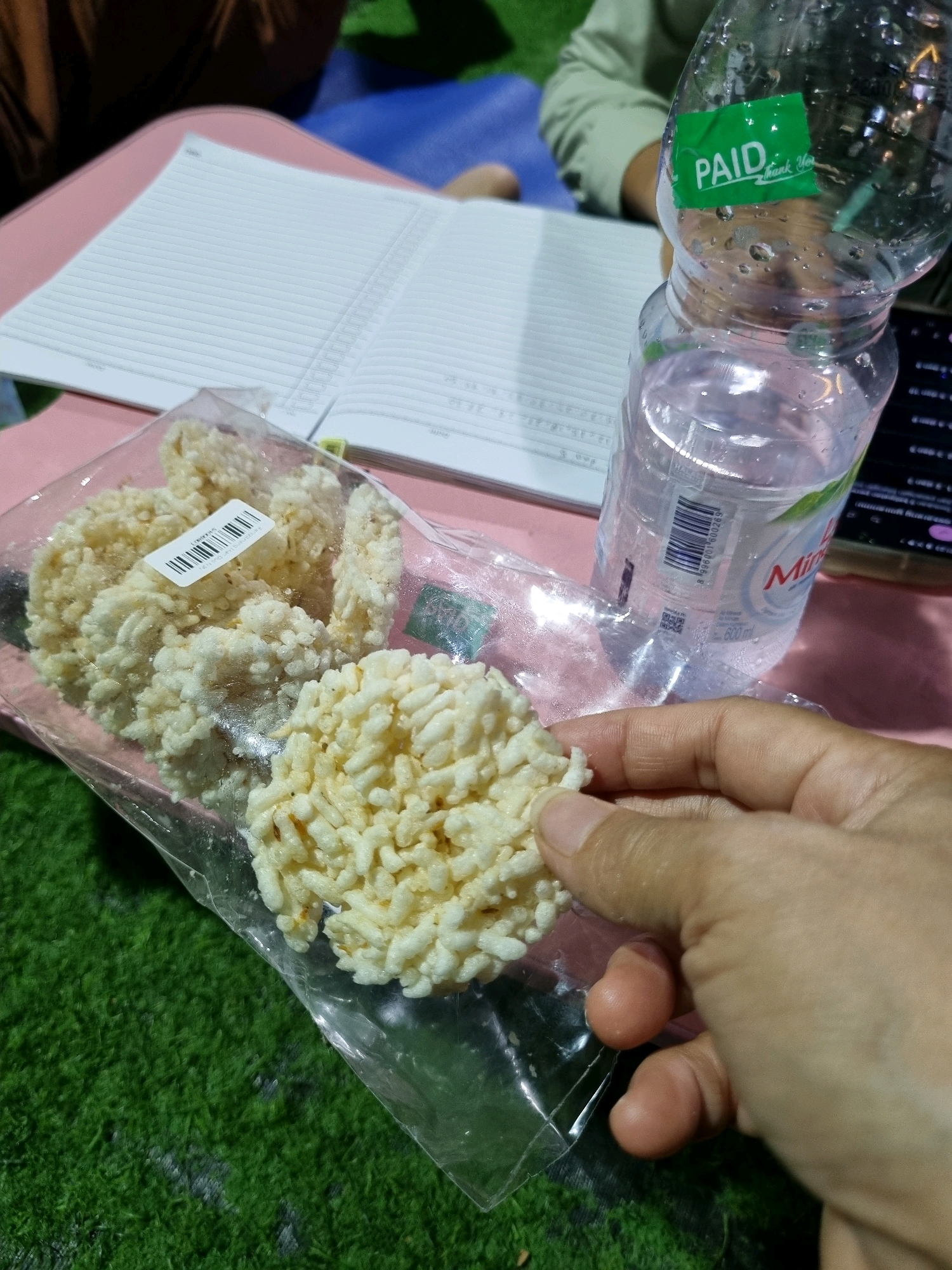 In Tanamodindi, Indonesia, this close-up, eye-level shot captures a hand holding a piece of puffed rice snack from a clear plastic bag. Behind the snack, a partially consumed bottle of water, labeled "Mines," sits on a pink surface, next to an open notebook. The background features a patch of green artificial grass and a blue surface, with hints of individuals in the upper portion of the frame. The scene appears to be outdoors or in a setting designed to mimic an outdoor environment.