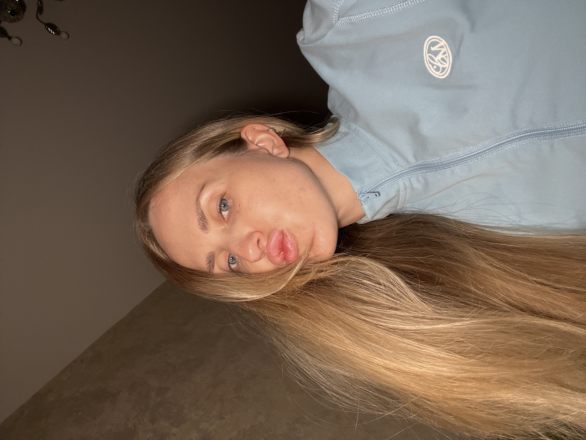 A woman with long blonde hair and blue eyes is captured in a close-up, low-angle shot. She is wearing a light blue zippered jacket with a circular logo on the chest. Her lips are pursed in a kiss. The background appears to be an indoor setting with textured walls and a ceiling. The lighting is dim, suggesting it might be evening or a low-light environment. The exact location within Unknown, Russia is not discernible from the image.