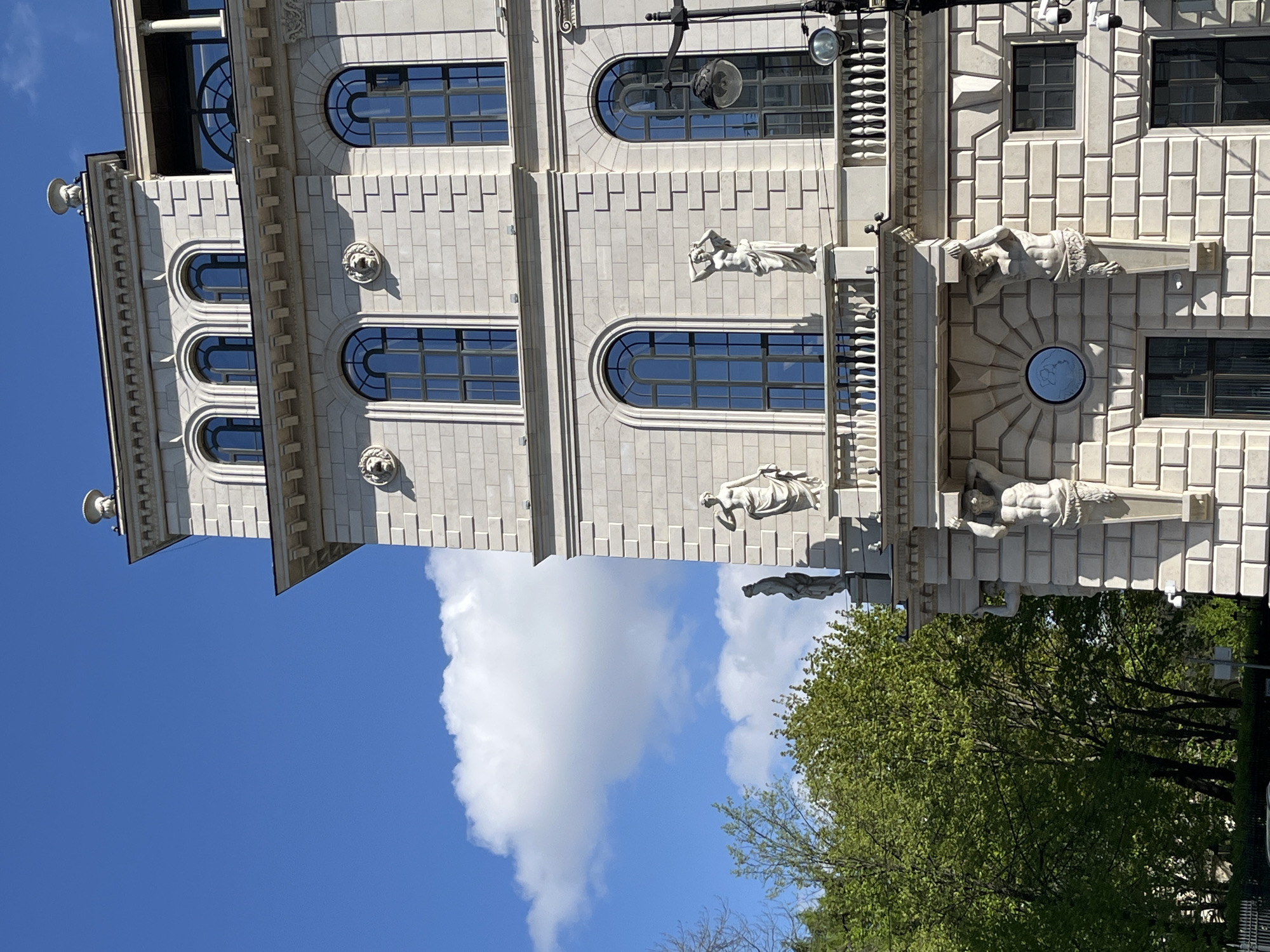 The image shows a building in St. Petersburg, Russia. The architectural style is neoclassical, with arched windows, decorative stone facades, and statues of figures supporting the building's cornice. A sky with some clouds is visible in the background. There are green trees in the lower right of the image.