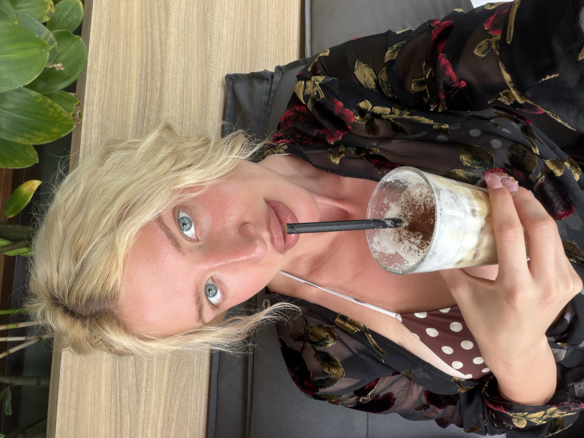 A young woman with blonde hair and blue eyes is holding a glass of iced coffee in her right hand, the glass topped with whipped cream and cinnamon. She is wearing a brown and white polka dot bikini top and a sheer black robe with a floral pattern. The woman is seated on a dark gray sofa, with a wooden surface and green plants visible in the background. The scene appears to be an outdoor setting, possibly a cafe or resort, in North Kuta, Indonesia.