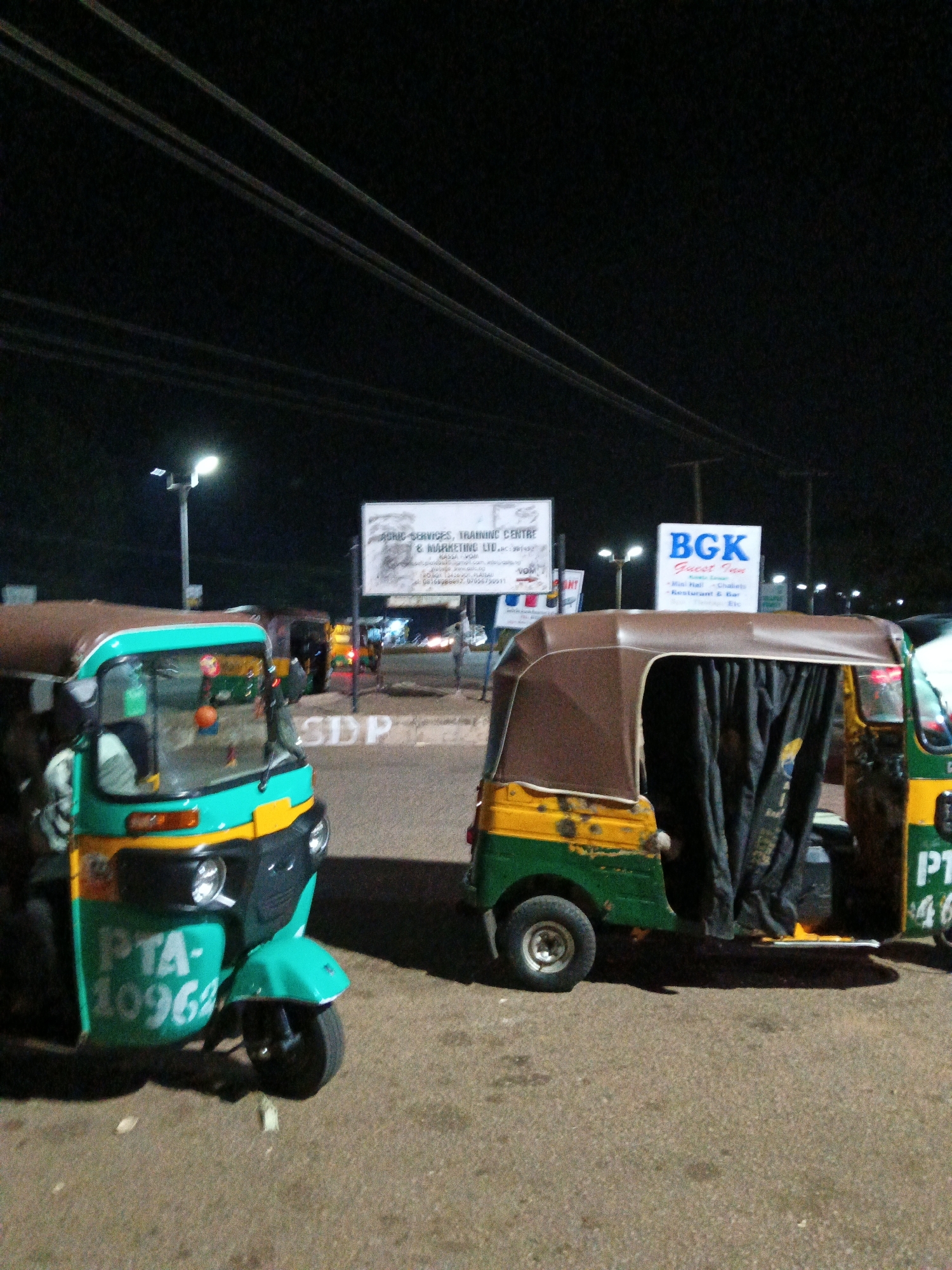 A nighttime outdoor scene in Bukuru, Nigeria, shows two auto-rickshaws parked on a paved surface. The rickshaw on the left is teal and yellow with "PTA-10962" written on its side. The rickshaw on the right is green and yellow, with a brown canvas cover over its passenger area. In the background, several signs are visible, including one that reads "BGK Guest Inn" and another for "AGRIC SERVICES, TRAINING CENTRE & MARKETING LTD." Streetlights illuminate the scene, and power lines are visible overhead. A few indistinct figures can be seen in the background, suggesting ongoing activity. The ground is a rough asphalt or concrete surface.