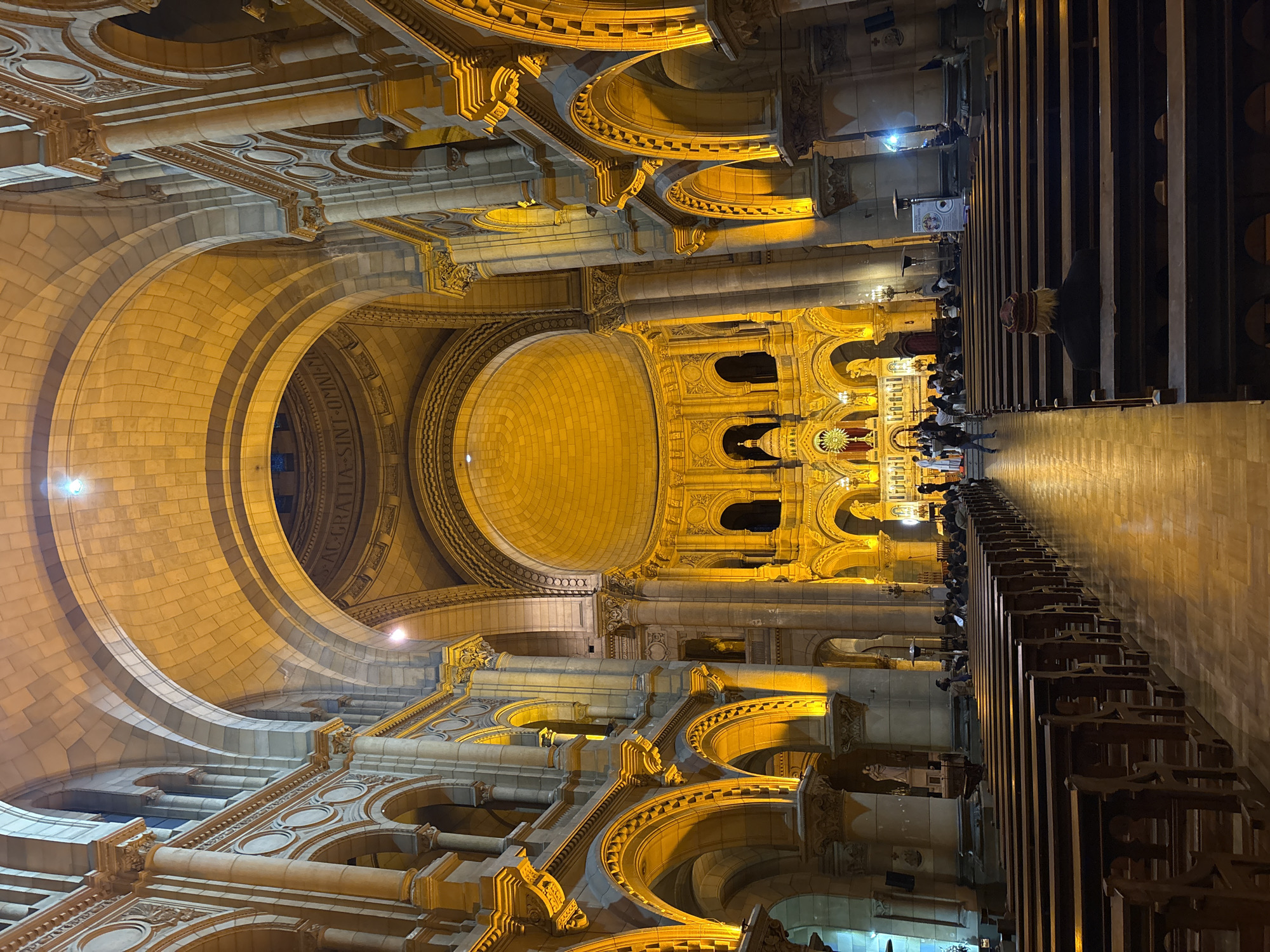 This is an interior shot of the Basílica de la Gratitud Nacional in Santiago, Chile, at night. The church is illuminated by warm, golden lights that highlight the stone architecture, including grand arches, columns, and a large dome. Rows of wooden pews fill the central nave, with some people seated in them. In the foreground, the back of a person wearing a knitted cap is visible, seated in a pew. Further down the nave, other individuals are seen walking and standing. The altar area at the far end of the church is brightly lit and appears to be decorated. The flooring is polished wood, reflecting the ambient light.