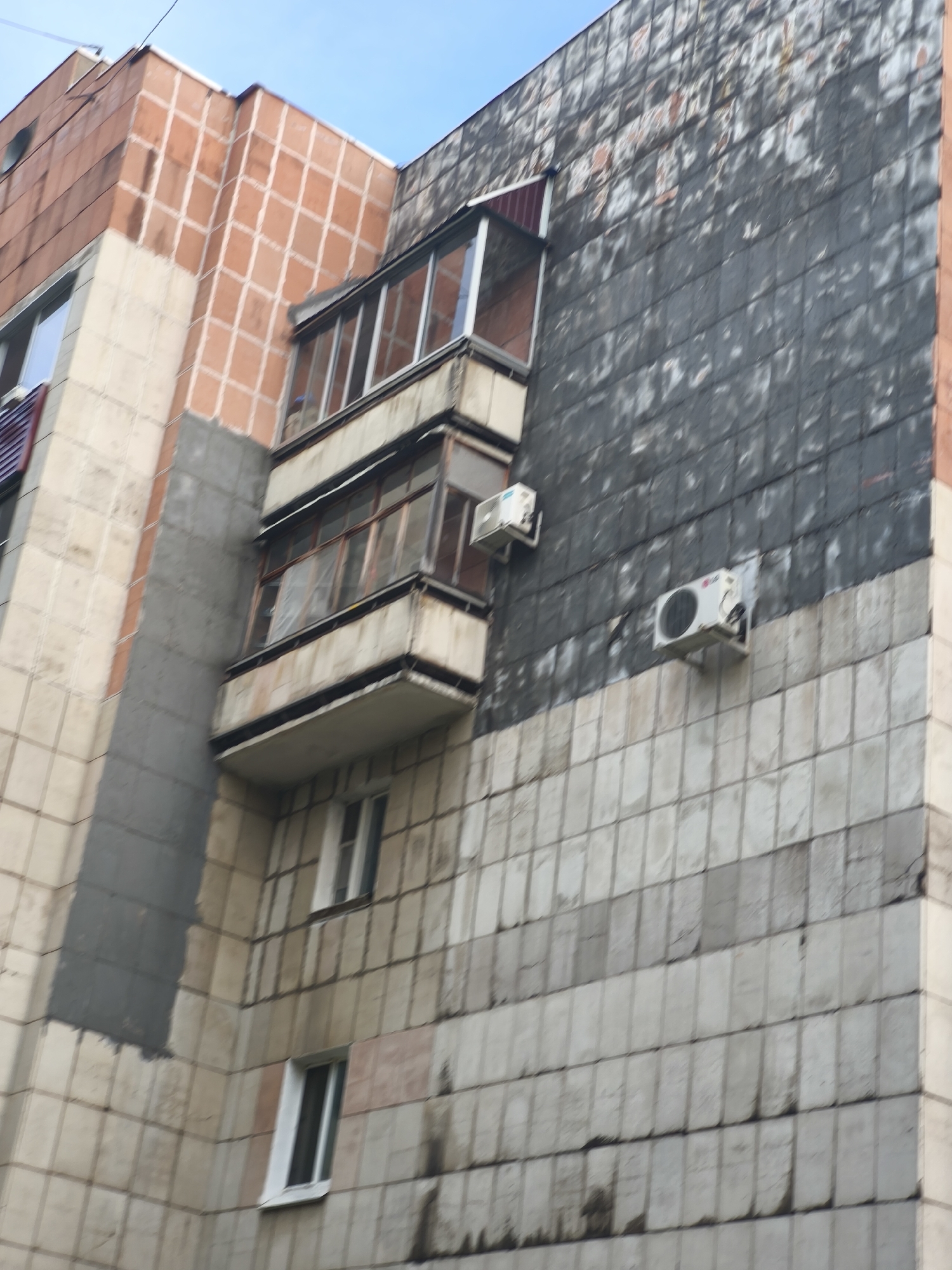 The image shows the exterior of a multi-story building, possibly an apartment building, with a mix of architectural styles and materials. The building features sections clad in light-colored tiles and a darker, possibly weathered, tiled section. There is a prominent balcony or enclosed porch extending from the building, with multiple panes of glass. Two air conditioning units are mounted on the exterior wall, one higher and to the left, and another lower and more centrally located. Windows of various sizes are visible, some with white frames. The sky is visible at the top of the image, appearing overcast or cloudy. The building appears to be in an urban environment, characteristic of Komsomolsk-on-Amur, Russia.