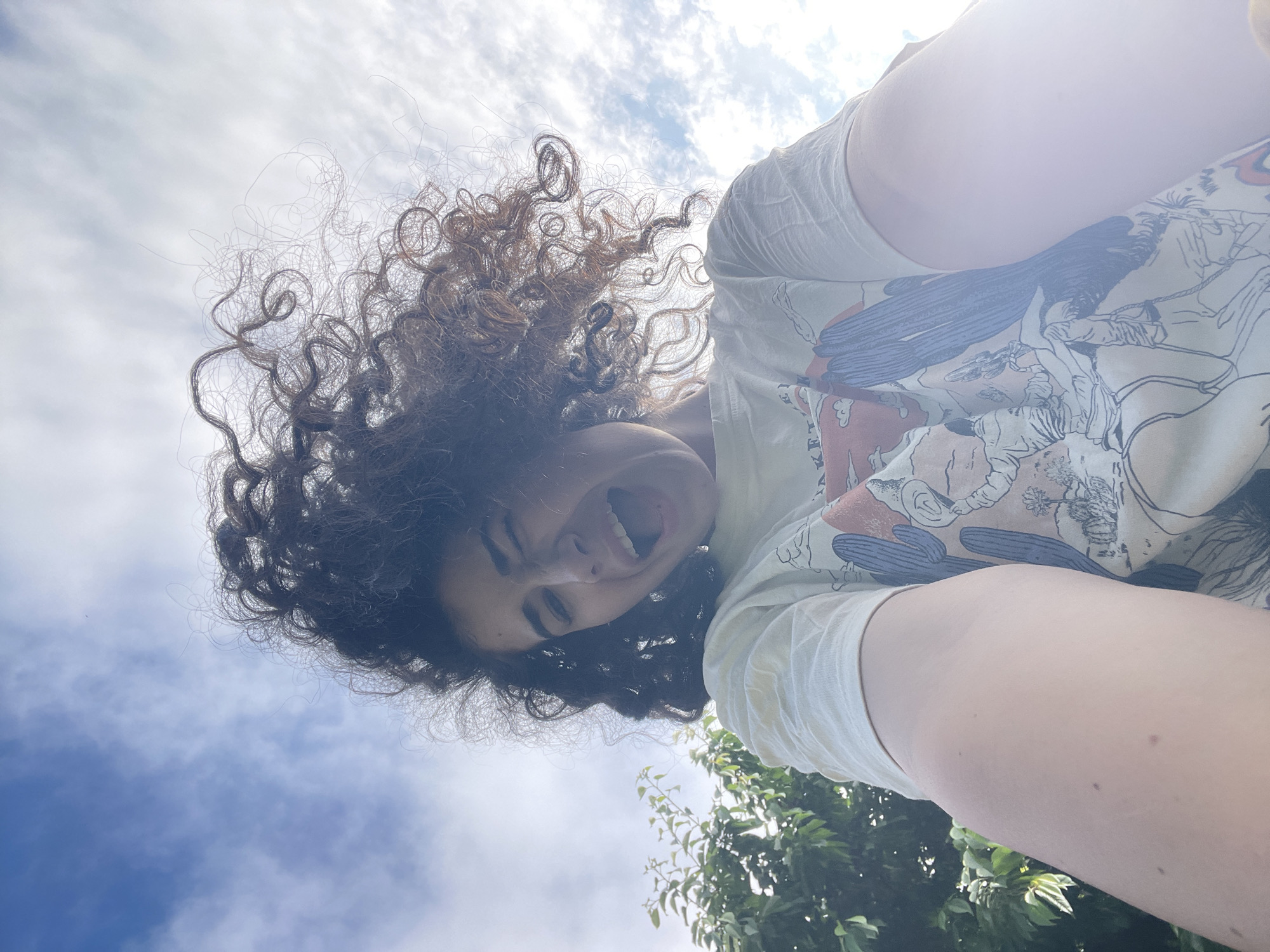A person with voluminous, dark, curly hair is captured in a low-angle selfie, looking down at the camera with a wide, joyful smile or laugh. Their head is tilted, and their abundant hair frames their face, spreading out against a bright, partially cloudy sky. Strong sunlight, causing a prominent lens flare in the upper right, indicates it is daytime.
The person wears a light-colored, possibly off-white, t-shirt adorned with a graphic design featuring blue cacti and other stylized figures, suggesting a desert or western theme. Green foliage from a tree or bush is visible along the bottom and right edges of the frame. The perspective creates an upward-looking view towards the person, emphasizing the vastness of the sky and the brightness of the day. The scene conveys a playful and happy mood. The image was captured in Sofia, Bulgaria. No clearly legible text is visible.
