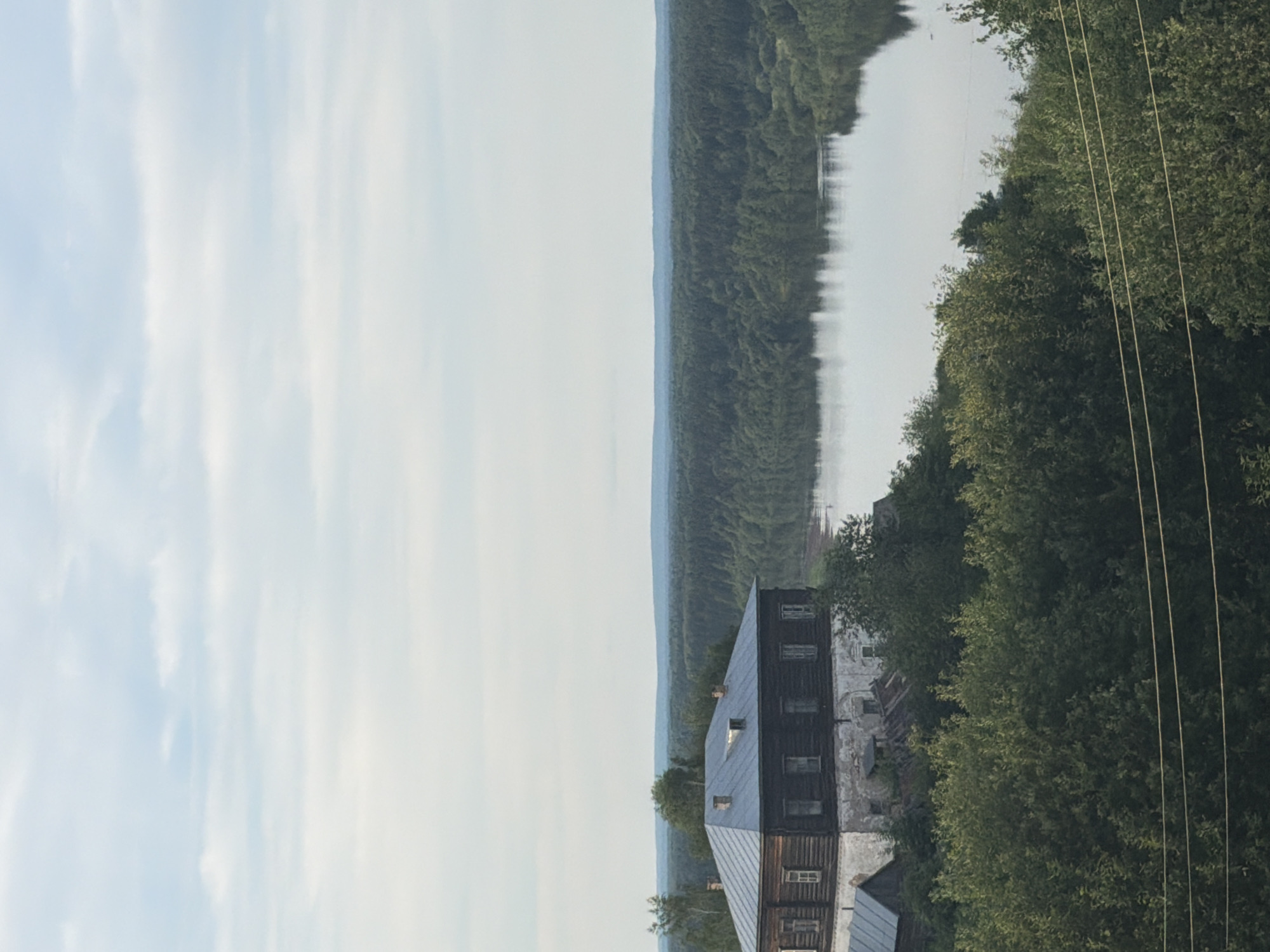 The image depicts a pastoral scene with a prominent wooden building, likely a traditional Russian house or barn, situated on the edge of a tranquil river. The building features a dark, weathered wood exterior and a sloping metal roof with visible windows. Adjacent to the building, lush green trees form a dense backdrop, extending towards the riverbank. The river itself appears calm, reflecting the overcast sky and the surrounding foliage. In the distance, a hazy line of hills or mountains can be seen, shrouded in a light mist, suggesting a cool or humid atmosphere. The sky is a soft, pale blue with streaks of white clouds, indicating it's likely daytime, possibly late afternoon or early morning due to the diffused light. The overall mood is serene and somewhat rustic, evoking a sense of quiet rural life. Power lines are visible in the foreground, crossing diagonally across the frame, hinting at the presence of modern infrastructure in this otherwise traditional setting. The image appears to be taken from a slightly elevated position, offering a panoramic view of the landscape. No people are visible in the scene, and there is no visible text.
