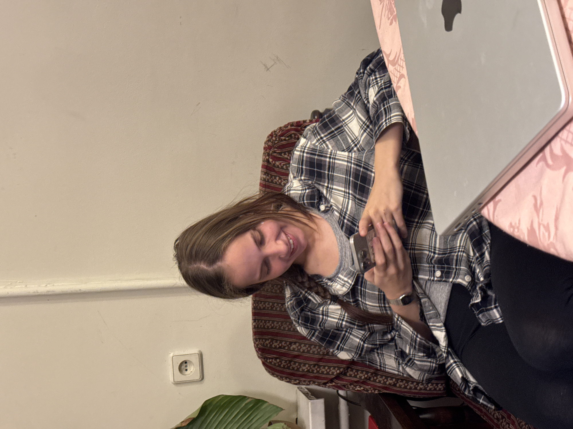 A young woman sits in a patterned chair, holding a smartphone. She wears a gray t-shirt under a blue and white checkered flannel shirt. She is wearing black leggings. A silver laptop sits on a pink patterned table to her right. The background includes a cream-colored wall with an electrical outlet and part of a plant. The location is not discernible as Moscow, Russia, from the provided visual information.