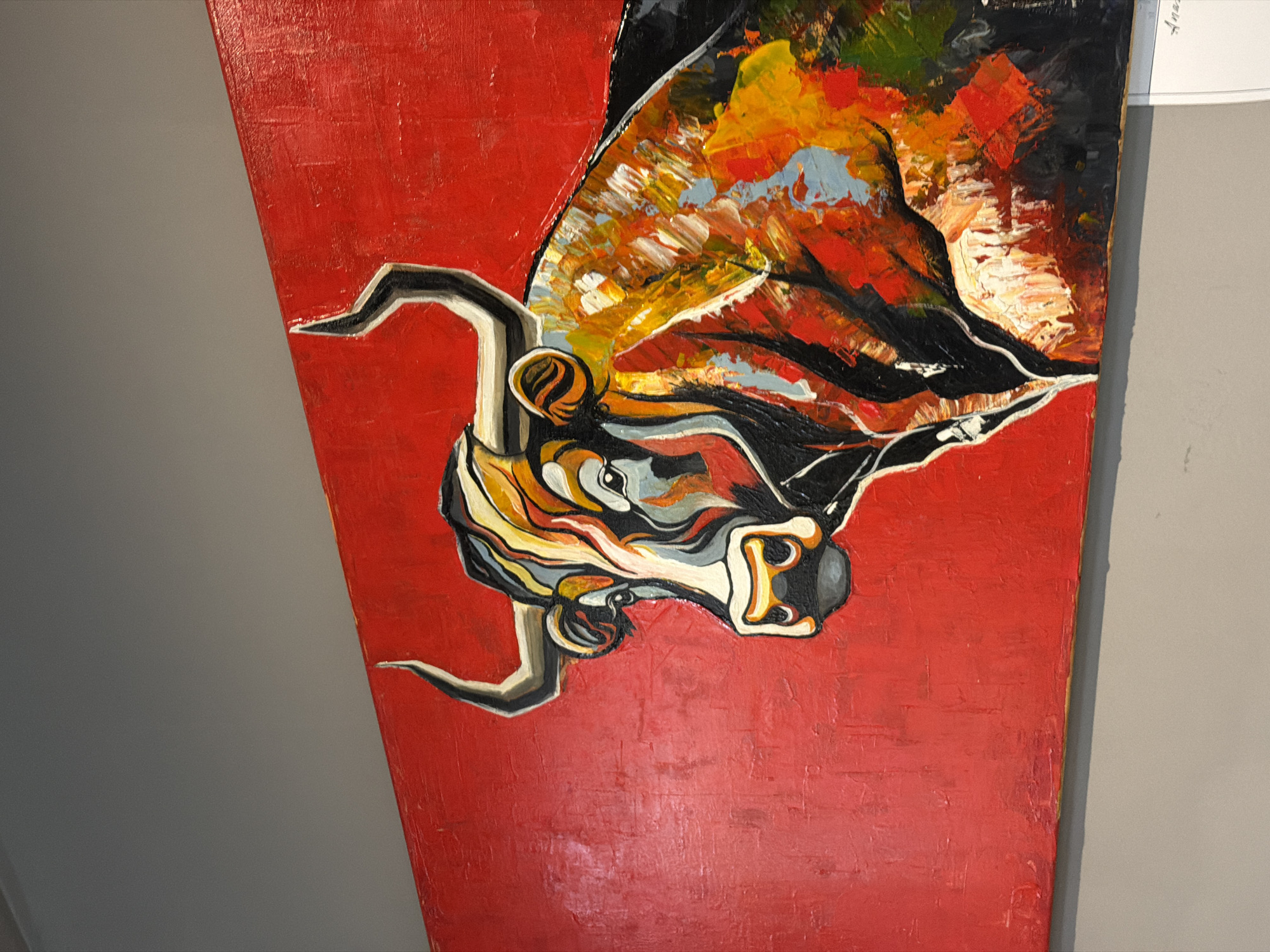 The image depicts a painting of a bull's head on a red background. The bull's head is painted in a stylized manner with thick brushstrokes of black, yellow, white, grey, and orange. The painting appears to be on a rectangular canvas. The environment is unknown. The city is unknown.