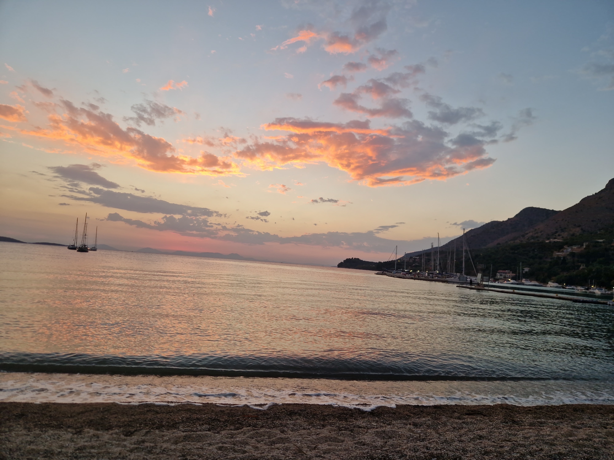 This image captures a serene sunset over the water in Plataria, Greece. The sky is painted with hues of orange, pink, and blue, with scattered clouds reflecting the last rays of the sun. Several sailboats are anchored in the calm water, their masts silhouetted against the twilight sky. In the distance, a coastline with hills and scattered buildings can be seen. The foreground features a pebble beach with gentle waves lapping at the shore, creating a peaceful and tranquil atmosphere. The lighting suggests it is late evening, just as the sun is setting below the horizon.