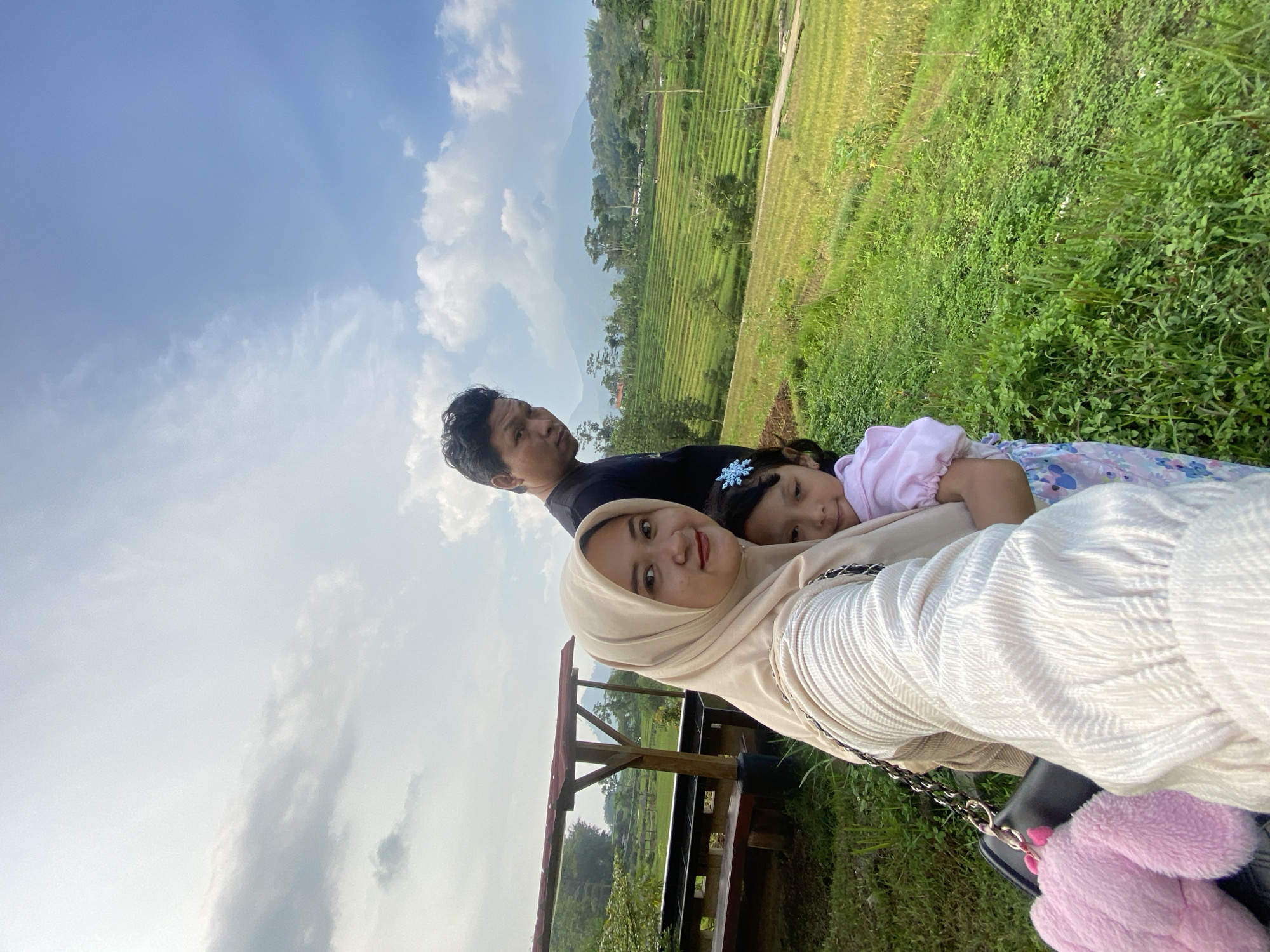 A family of three poses for a selfie at Penanggungan, Indonesia. The image is slightly tilted clockwise. A woman wearing a beige hijab and a white textured top is in the foreground, holding the camera. A man wearing a black shirt is slightly behind her, and a young girl wearing a pink top and a blue flower hairpin is nestled against the woman. They are on a wooden structure with a partial roof, and a green, lush landscape with terraced fields extends behind them. The sky is visible with white clouds and blue patches. A pink plush accessory hangs on a black bag.
