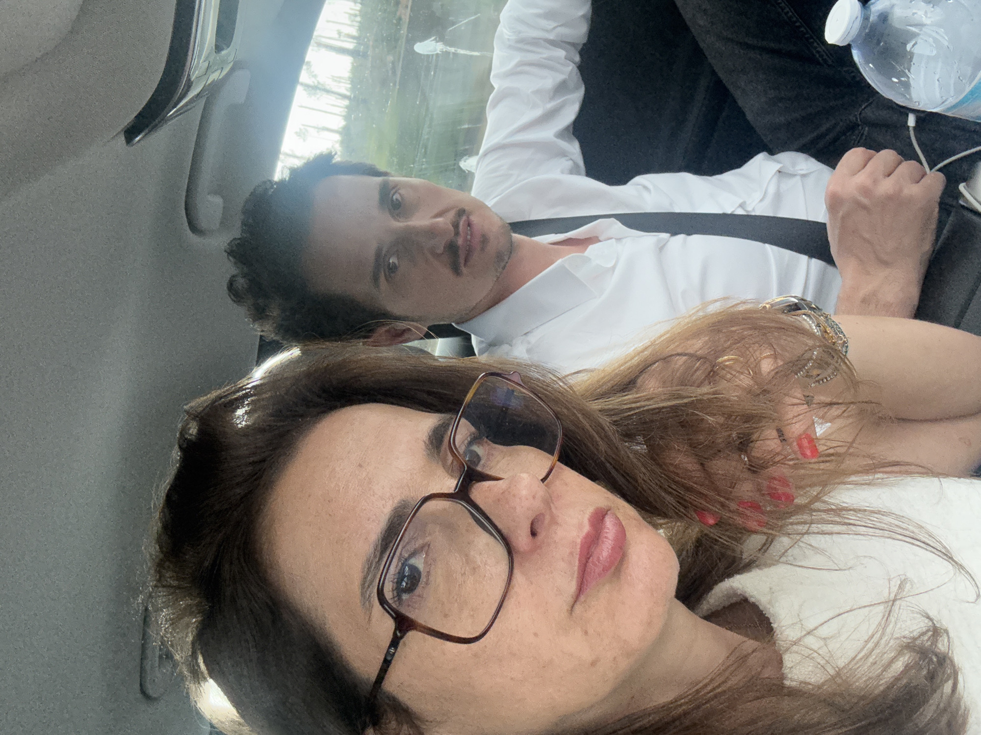 A selfie-style photograph shows two individuals inside a vehicle. The woman in the foreground wears glasses and a light-colored garment. Behind her, a man in a white shirt is partially visible, wearing a seatbelt. A bottle of water is present near the man. The background appears to show an exterior scene possibly in Alcobaça, Portugal. The car interior is visible with a headrest and handle. The image is slightly angled.