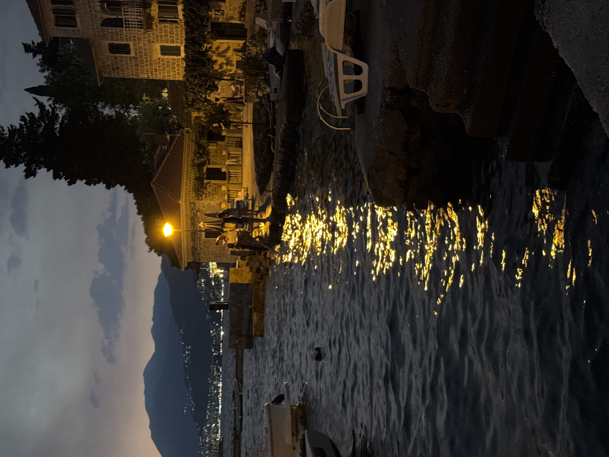 This is a nighttime, slightly low-angle, vertical shot of a waterfront area in an unspecified location. The foreground is dominated by dark, rippling water with a strong reflection of yellow light from an unseen source, likely streetlights or lamps, creating a shimmering effect on the surface. To the right, a concrete embankment rises from the water, with rough stone steps leading up to a flat surface. On this surface, a white lounge chair with a metallic frame is positioned, facing the water. Further back, a stone building with a tiled roof and arched windows is visible, partially obscured by dark foliage. Several people are gathered in front of this building, illuminated by artificial light, with some appearing to be standing and others seated. A yellow lamppost is also visible in this area. In the background, across the water, a distant city skyline with numerous lights is spread out along the base of dark, silhouetted mountains under a twilight sky. The water in the midground contains a small, dark object that appears to be a duck or a similar waterfowl. Several small boats are moored along a stone pier in the midground left.