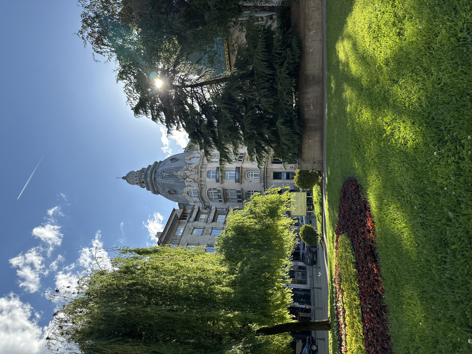 The image is an upward angle shot from ground level, showcasing a cityscape in Sofia, Bulgaria on a sunny day. An ornate, multi-story building with a prominent dome, likely a historical edifice, dominates the view, partly obscured by green trees. The sun, positioned in the upper part of the frame, adds a flare effect. A contrail cuts across the bright blue sky dotted with fluffy white clouds. To the right of the building and its partially obstructed view of green landscape, including a manicured lawn and flower bed. Below the building and to the left, green trees frame the lower portion of the image, partly hiding parked cars and the façade of buildings along the sidewalk.