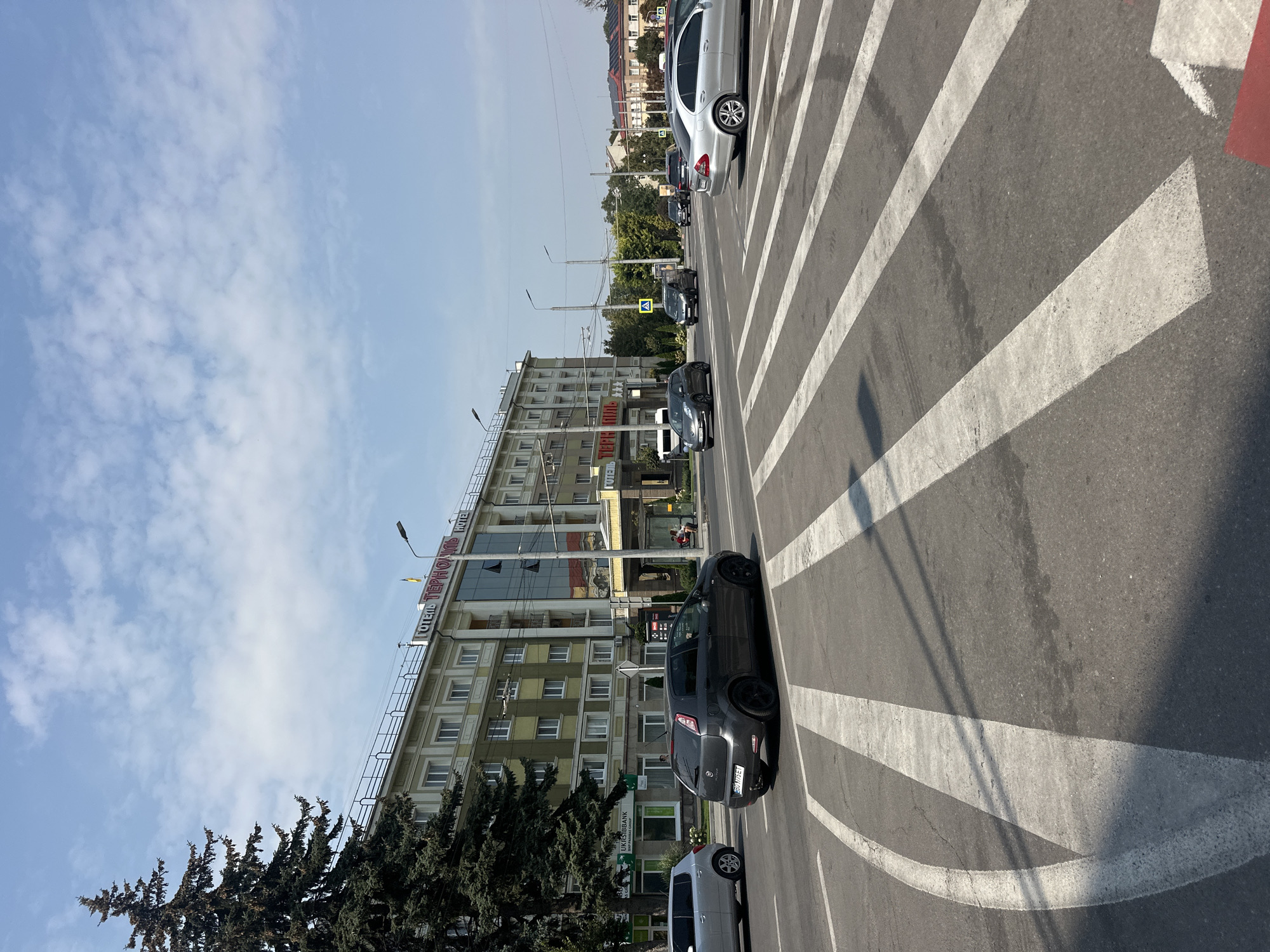 The image shows a street scene in Ternopil, Ukraine. In the foreground, a crosswalk is marked with white stripes on asphalt. Several cars are visible on the road. A dark gray Fiat Punto is in the left lane, and a silver Mercedes-Benz sedan is in the right lane. Further down the road, a dark SUV and other vehicles are present.
On the left side of the street, a large evergreen tree stands next to a multi-story building. This building appears to be a hotel, with "ГОТЕЛЬ ТЕРНОПІЛЬ HOTEL" (Hotel Ternopil Hotel) signage visible near the top. The building has a light green facade with rows of windows.
On the right side of the street, more buildings are visible, including a structure with a red roof. The sky is blue with scattered white clouds. Streetlights and traffic signals are also present. The overall impression is of a moderately busy urban environment on a clear day.