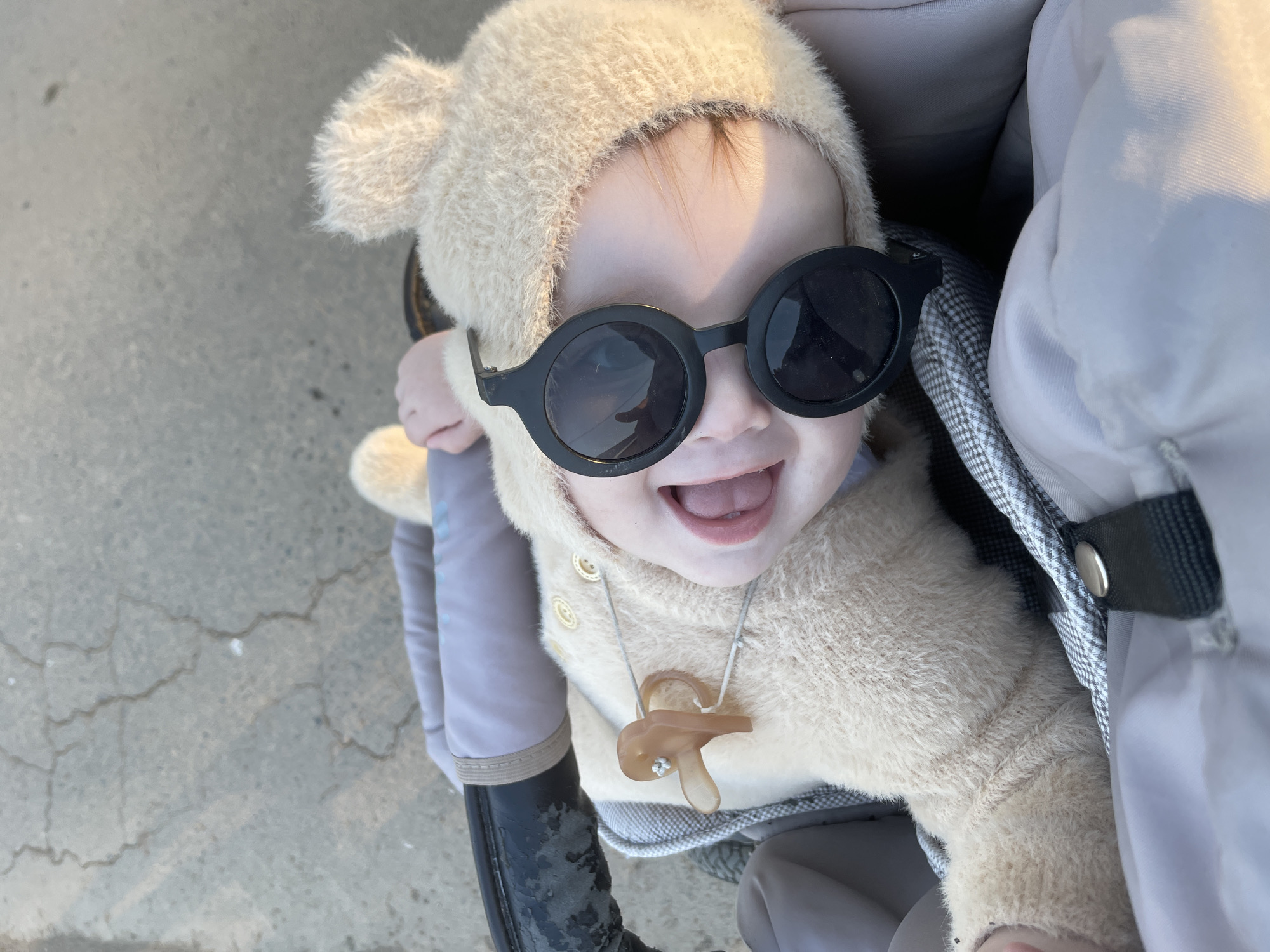 A baby wearing a furry bear-like hooded jacket and oversized round black sunglasses is seated in a stroller. The baby is smiling, revealing its teeth. A light brown pacifier on a string is around the baby's neck. The stroller appears to be on a paved surface. There's no specific indication it is Подшивалово, Russia.