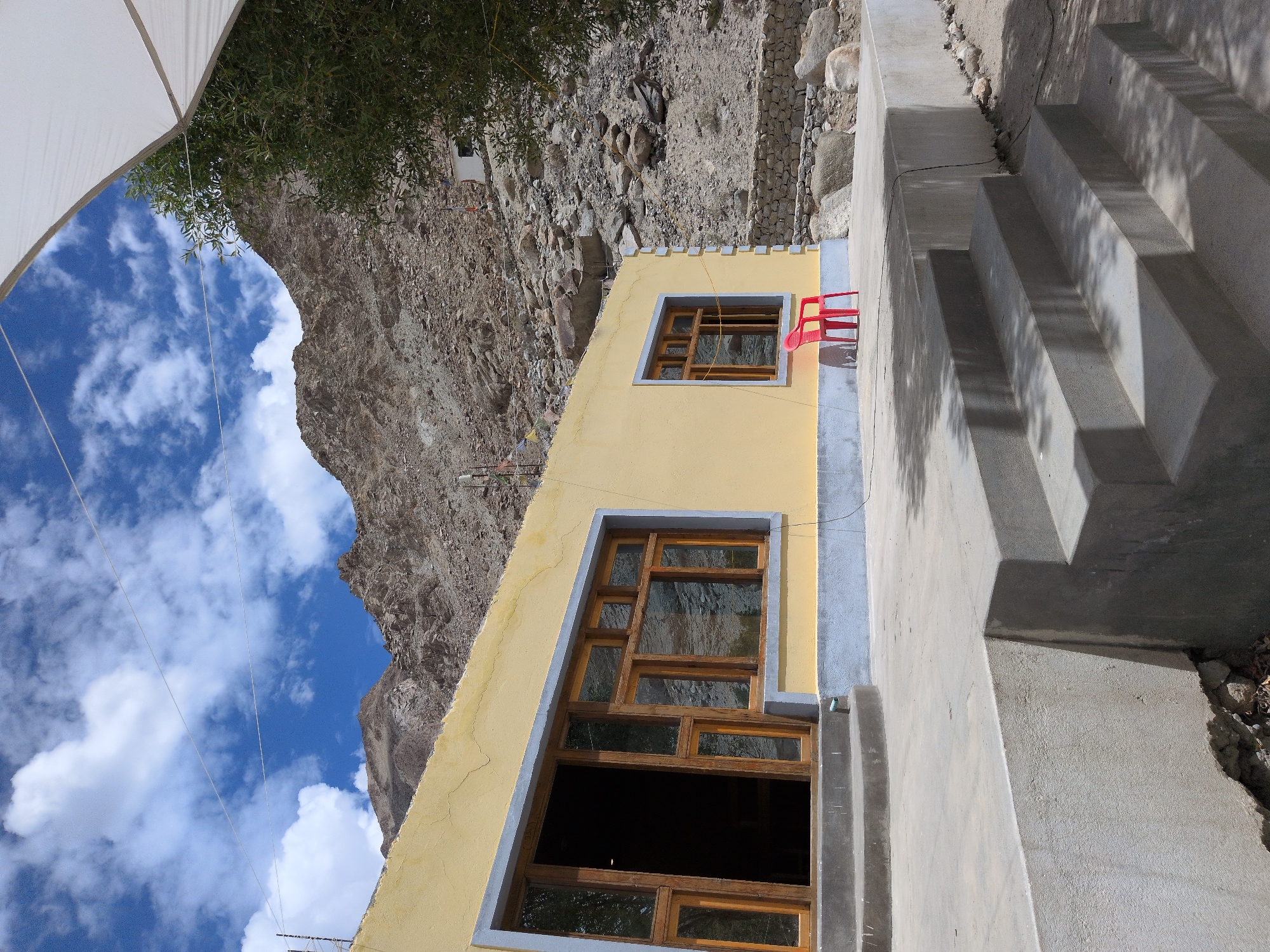 The image shows a yellow building with wooden framed windows and doors, situated in a mountainous, arid environment. A red plastic chair is placed on the concrete patio in front of one of the windows. Steps lead down from the patio to a sandy ground. The background features a rocky, barren mountain slope with scattered boulders and sparse vegetation. Prayer flags are visible on the hillside, suggesting a spiritual or cultural significance to the location. The sky is bright blue with scattered white clouds. The scene appears to be in Panamik, India, a region known for its natural hot springs and Buddhist monasteries.
