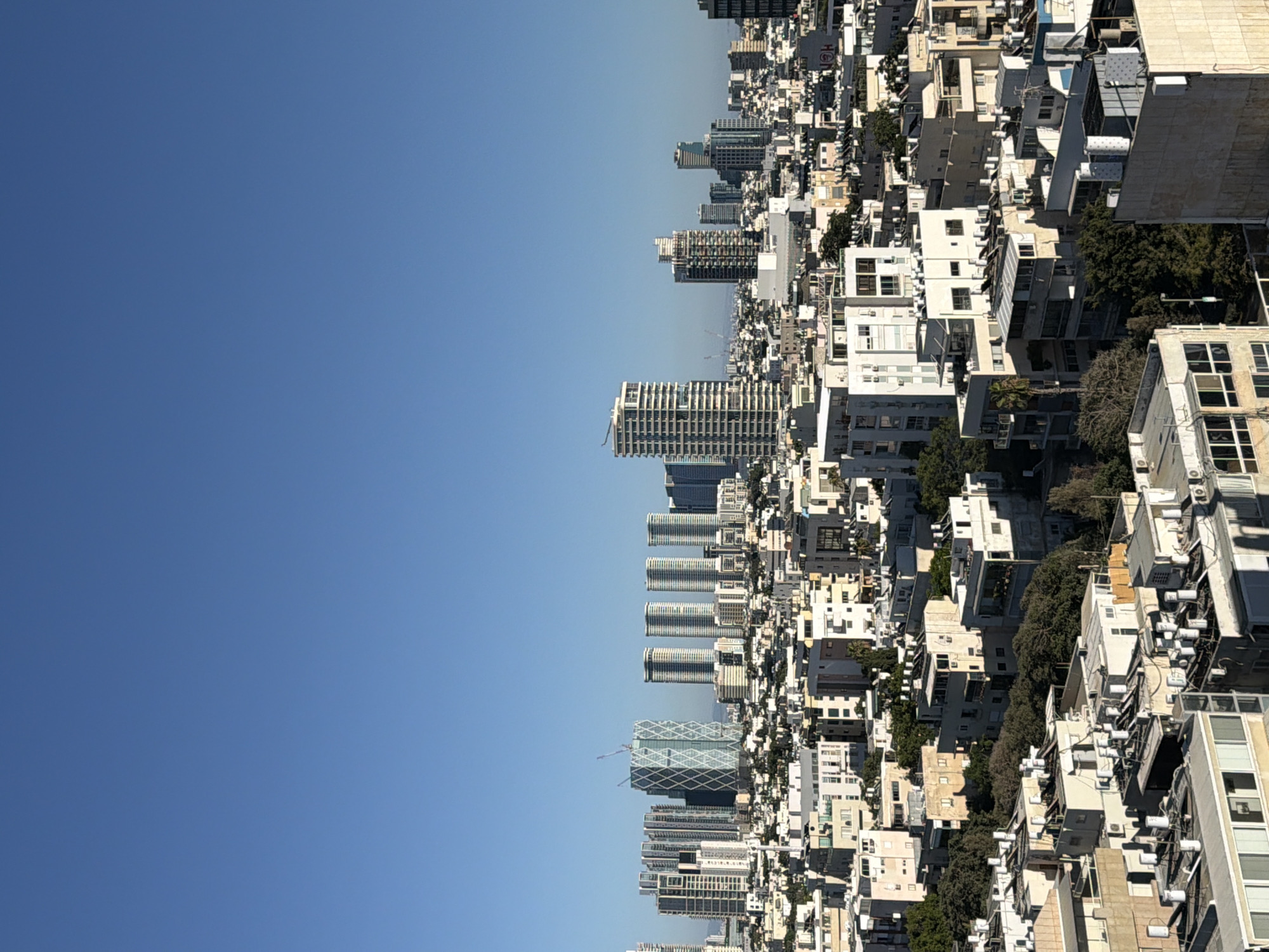 The image shows a panoramic view of the Tel Aviv skyline under a clear blue sky. The city is densely populated with a mix of modern high-rise buildings and lower-rise residential structures. In the background, several prominent skyscrapers stand out, including a distinctive diamond-patterned building that appears to be under construction. The foreground consists of a more densely packed urban landscape with buildings of varying heights and architectural styles. Many buildings feature flat roofs with numerous solar water heaters, a common sight in Israeli cities. Lush green trees are interspersed among the buildings, indicating a well-established urban environment. The overall impression is one of a vibrant and modern city with ongoing development. The image appears to be taken from an elevated position, offering a broad perspective of the urban expanse. No people are visible in the image.