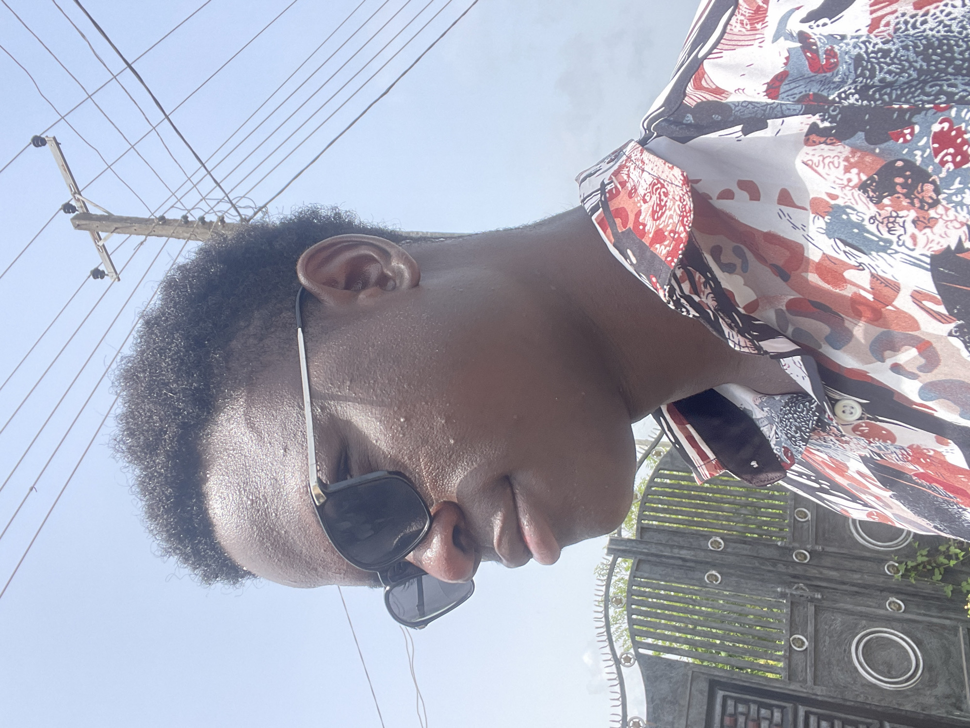 A low-angle, medium shot of a person with dark skin and short, curly hair wearing sunglasses. They are standing near a dark gray metal gate with decorative circular elements. The person is wearing a patterned shirt with reds, whites, grays, and blacks. In the background is a clear blue sky and telephone poles with wires. The location is likely Badagry, Nigeria, judging by context.