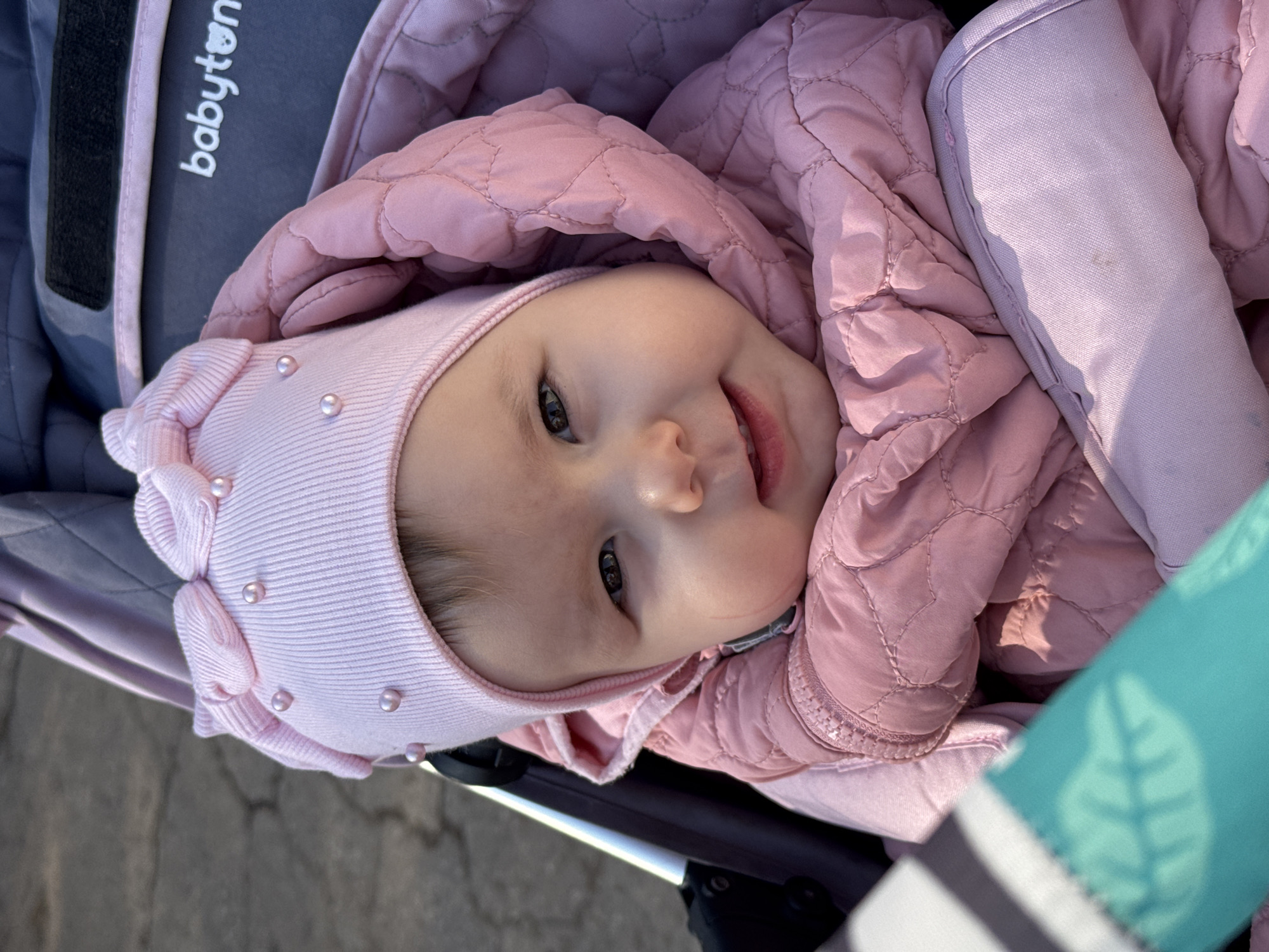 A close-up, angled, outdoor shot shows a baby lying in a stroller. The baby has fair skin, brown hair under a light pink knit hat adorned with small pearls, and is smiling slightly. The baby is wearing a light pink quilted jacket. The stroller is visible in the frame, with parts in grey and light pink. "babyton" is printed on the side of the stroller. Part of the pavement is visible in the lower left corner. The lower right corner shows a turquoise-colored stroller part. The location is Zav'yalovo, Russia.