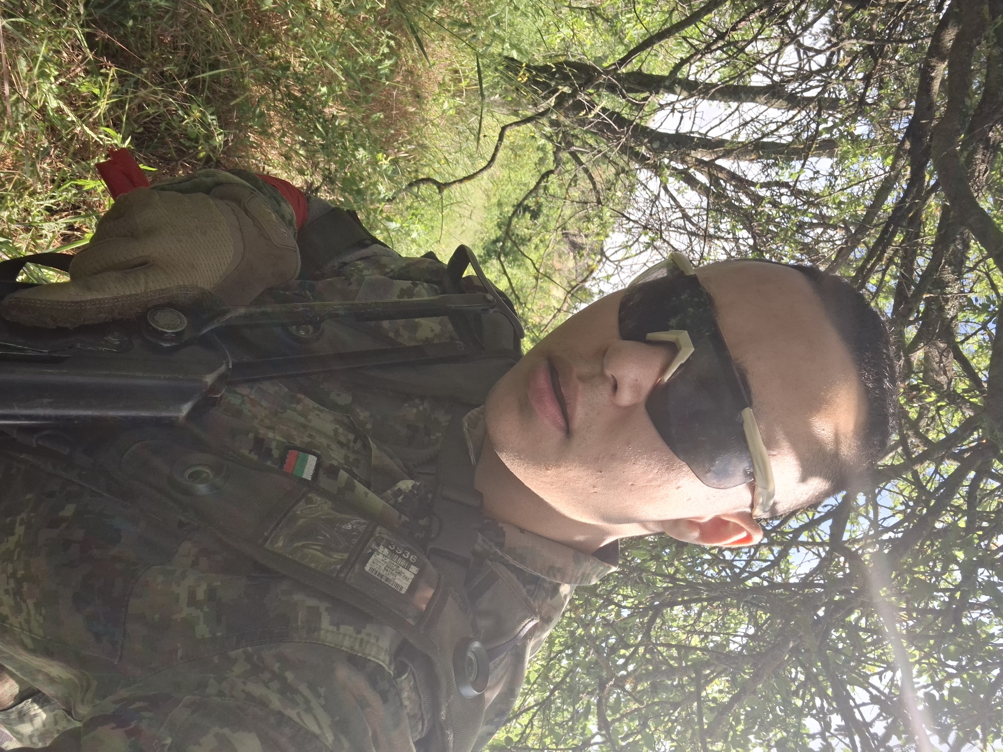 In a close-up, outdoor, selfie-style photograph, a person wearing camouflage tactical gear is shown from the chest up. They have dark, short hair and are wearing dark sunglasses. The person is outdoors, surrounded by lush green foliage and trees, with sunlight filtering through the branches. The background suggests a natural, wooded environment. The person is holding what appears to be a firearm, and they have a flag patch on their uniform, which appears to be the Bulgarian flag. The overall scene suggests the person is in a natural outdoor setting, possibly engaged in an activity like airsoft or military training, given the attire and equipment.