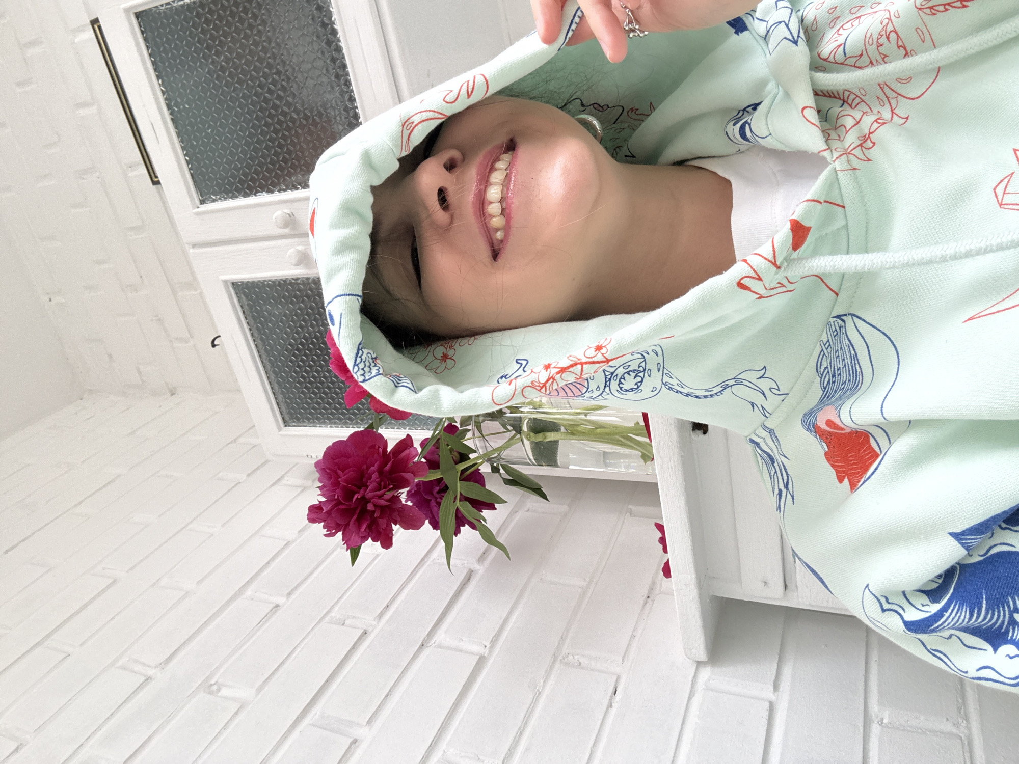 The image shows a young woman lying down, her head and shoulders covered by a light green hooded sweatshirt with a red and blue pattern. She is smiling with her eyes closed and her teeth visible. A few dark pink flowers in a clear glass vase are placed on a white wooden shelf near her. The background is a white brick wall and a white cabinet with frosted glass doors. The lighting suggests it is daytime. The woman is wearing a ring on her right index finger. There is no visible text in the image.