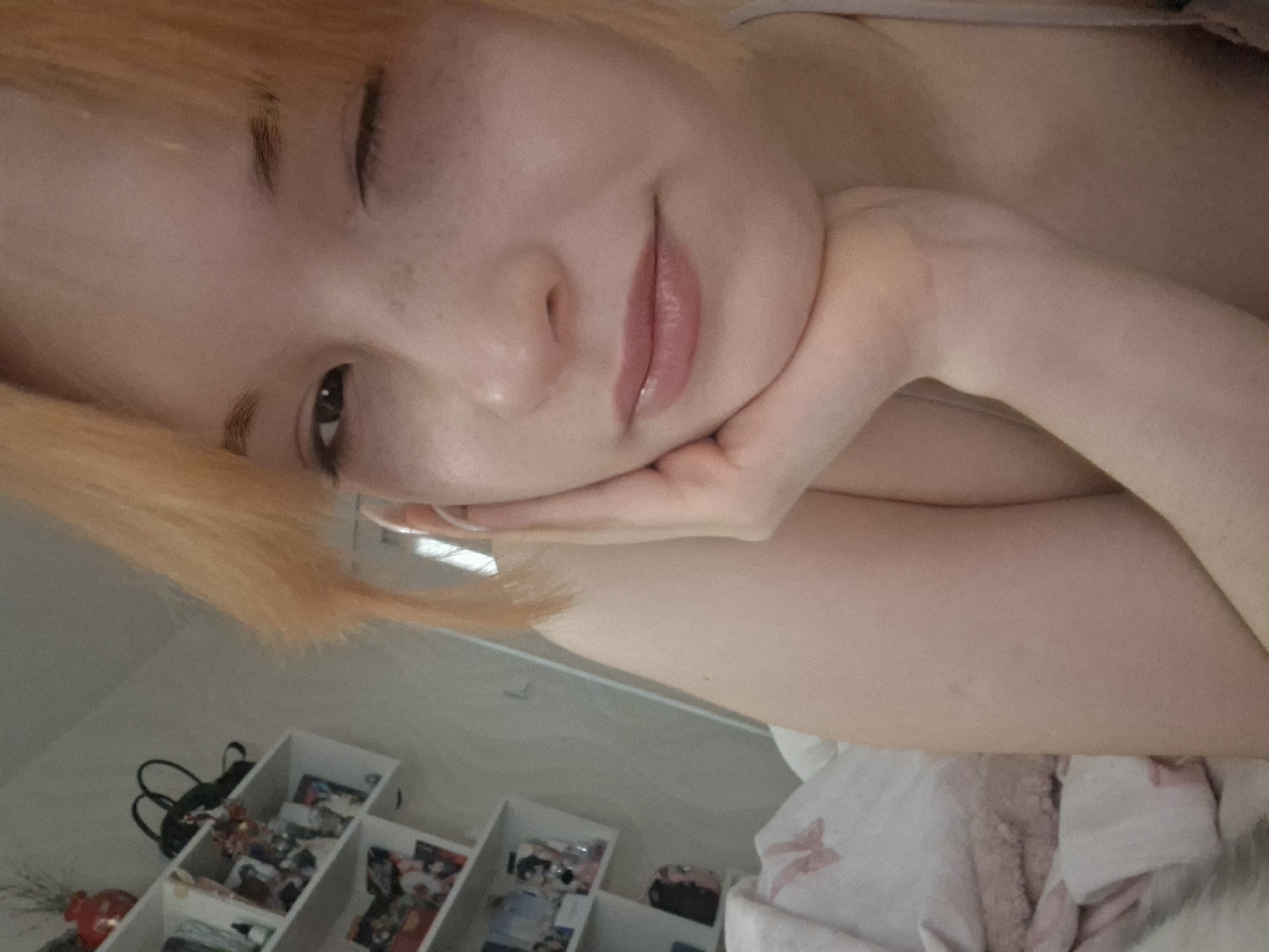 A selfie-style image depicts a fair-skinned person with short, orange hair. The individual rests their head on their hand, looking towards the camera with a slight smile. A set of shelves with various small objects are visible in the background, along with what appears to be bedding. There is no information to confirm that this image was taken in Chaikovsky, Russia, or any other location.