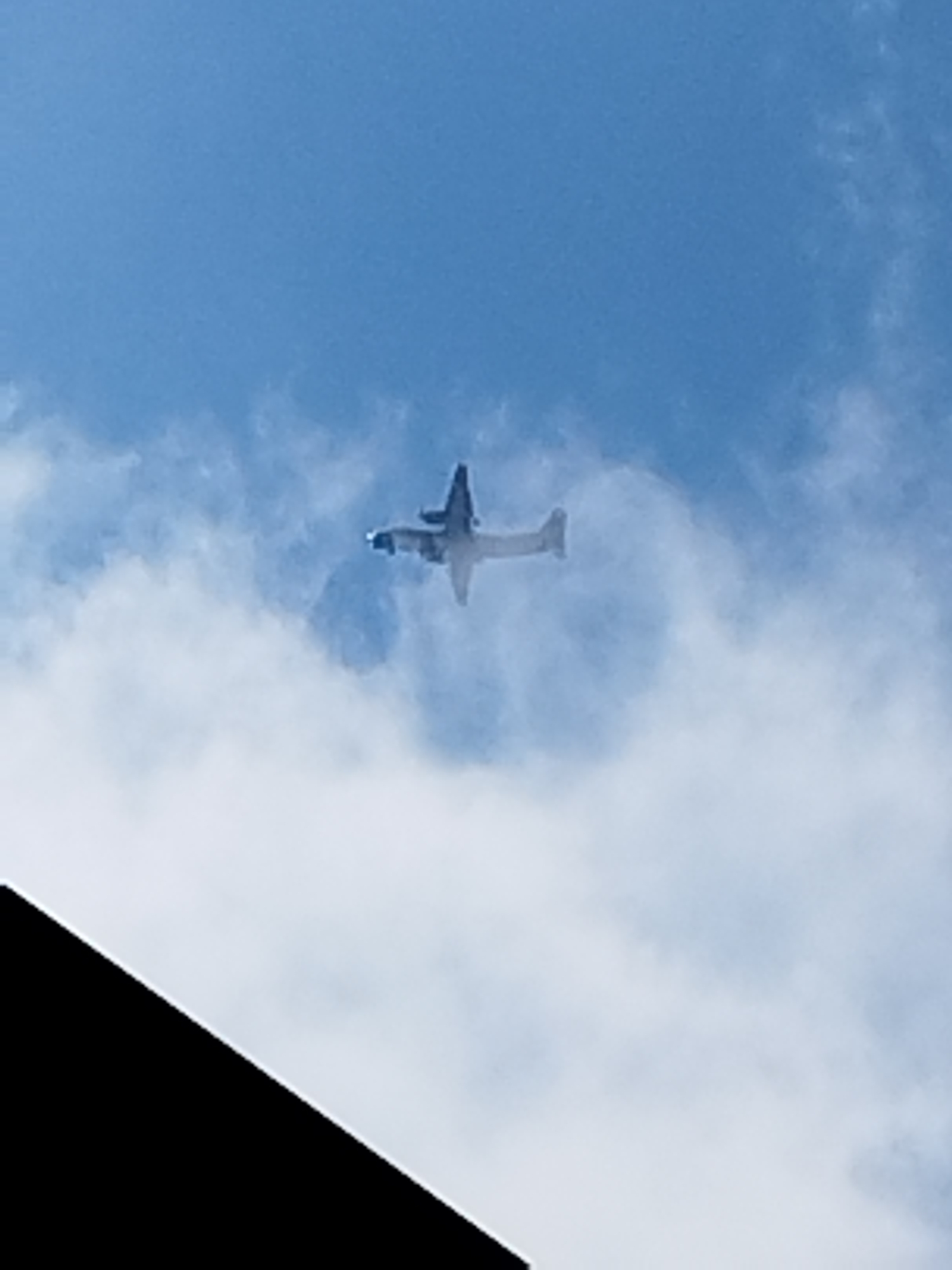 An airplane is seen flying in a cloudy sky. A dark, angled object, likely a building or structure, is in the lower left portion of the image. The image appears to have been taken from a low angle looking up at the sky. No specific location in Buftea, Romania is identifiable from the image.