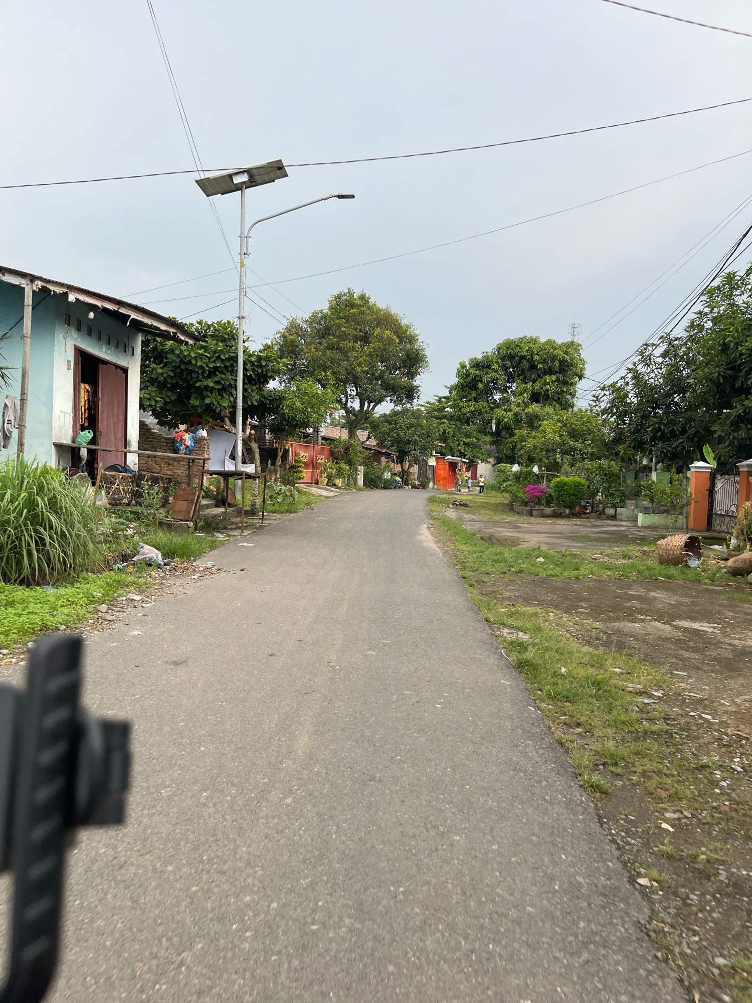 The image depicts a view down a paved street in Patumbak Kampong, Indonesia, likely on an overcast day. On the left side of the street, a light blue house with a brown-trimmed doorway is visible, with laundry hanging outside. Adjacent to the house is a solar-powered street lamp. Further down the street on the left, there are trees and foliage. On the right side of the street, there are houses with orange gates and fences, and manicured gardens with potted plants and flowers. The street itself is asphalt and appears to be a residential road. In the foreground, a dark, out-of-focus object, possibly part of a vehicle or camera mount, obstructs the lower left corner of the image. Several people, appearing as small figures, are visible in the distance on the right side of the road, near the orange gate. The overall environment appears to be a semi-urban or rural residential area.