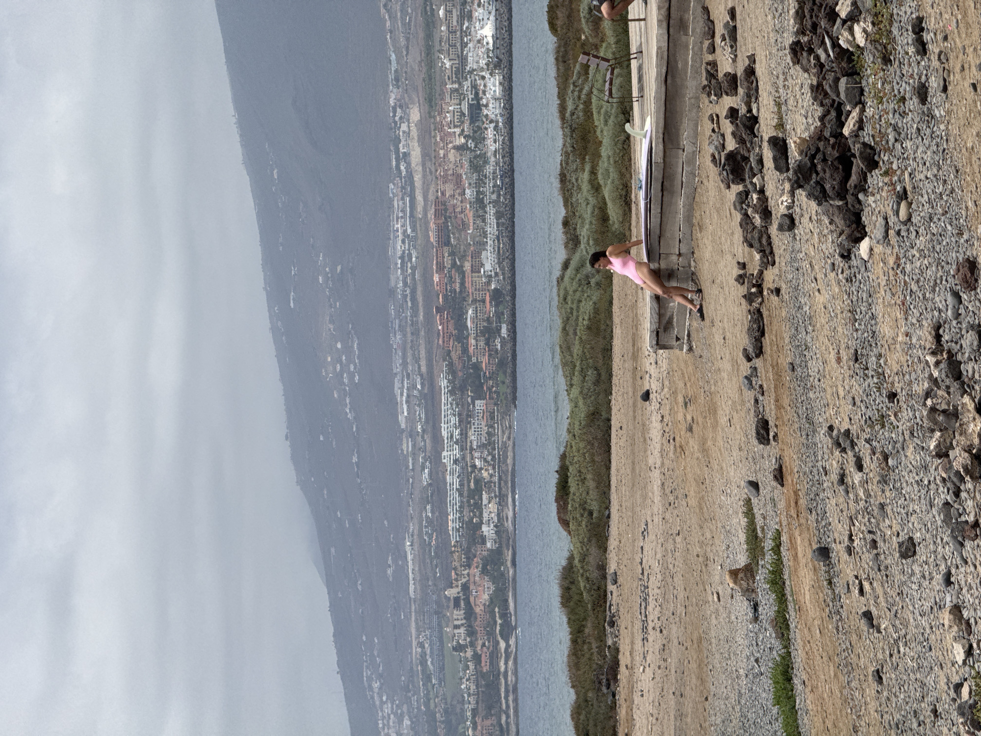 The image depicts a beach scene in Arona, Spain, under a cloudy sky. In the foreground, a rocky, sandy terrain is visible. A young woman, wearing a pink swimsuit, is seated on a low concrete structure with a surfboard resting beside her. She is looking towards the ocean. Behind her, a metal chair is placed on a wooden deck. In the middle ground, the blue ocean stretches out, with waves. Further back, a coastal town with numerous buildings is spread across a hillside, leading up to a mountain range in the distance. The overall atmosphere is muted due to the overcast weather.