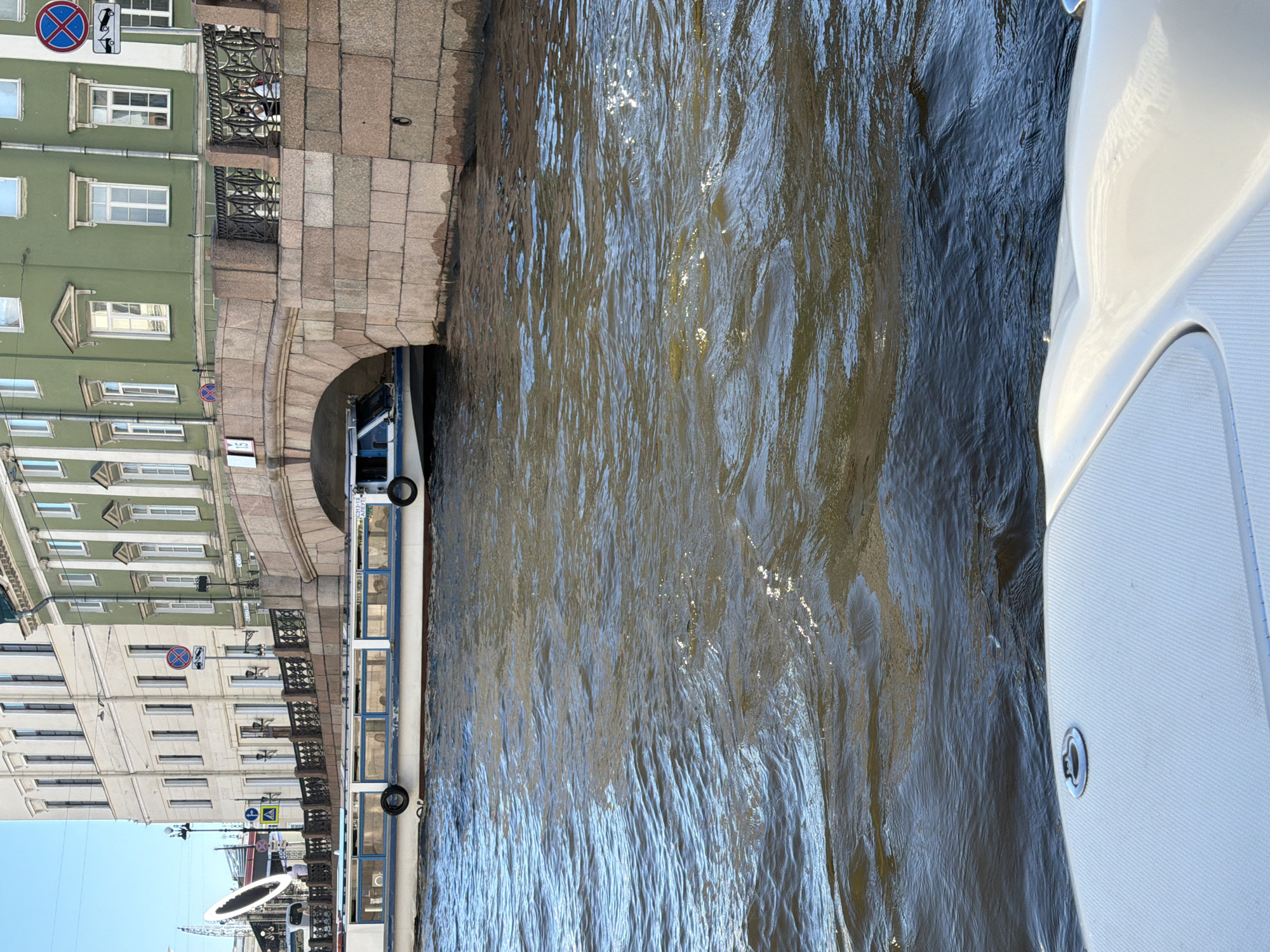 The image depicts a wide, brown waterway in the city of Saint Petersburg, Russia, flowing beneath a stone arch bridge. On the left, a white and blue passenger boat with large windows is visible, partially obscured by the bridge. The boat has black tires attached to its sides. The bridge is constructed of rough-hewn pink stones and features a black metal railing along its upper edge, above which are green and beige buildings with numerous windows. Signs are posted on the buildings and bridge. In the foreground, the white deck of a boat is visible, angled slightly towards the water. The water's surface is rippled, reflecting the sunlight and showing streaks of darker and lighter shades.