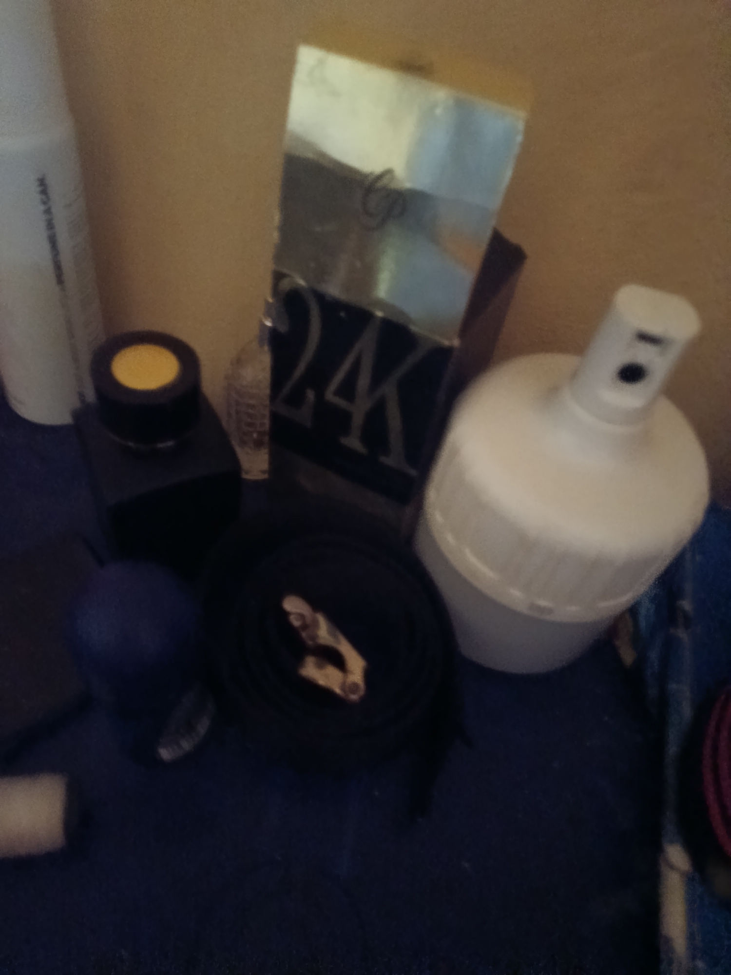 In the image, several objects are arranged on a dark blue surface, which is set against a tan background. On the left, a tall white bottle with text is visible. Next to it, a small black box with a yellow-capped lid sits. To the right of the black box, a partially obscured decorative glass bottle is present. Further to the right, a large, tall, rectangular object with "24K" prominently displayed in gold is seen. To the right of this, a white aerosol can with a black nozzle is situated. In the foreground, a dark coiled object with a gold clasp is visible. A dark blue spherical object is to the left of the coiled object. The overall scene appears to be a collection of cosmetic or personal care items. No people are present. The location is unknown.