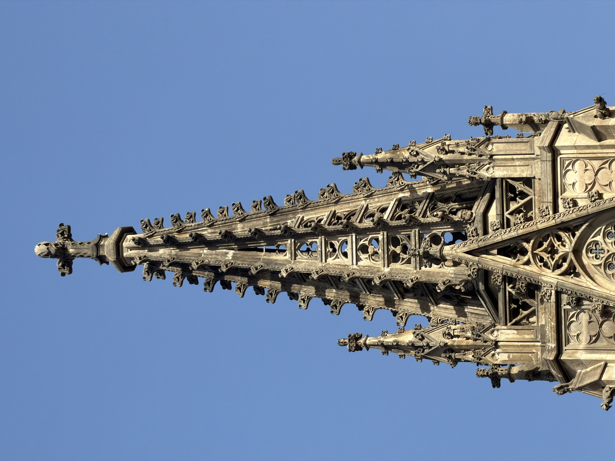 The image shows the elaborate spire of a Gothic cathedral, silhouetted against a clear blue sky. The spire is intricately decorated with numerous gargoyles, statues, and ornate stonework. At the very apex of the spire is a cross. The detailed architecture suggests a significant historical and religious structure. The image was taken from a low angle, emphasizing the height and grandeur of the spire.
This is a close-up, exterior, low-angle, eye-level shot of the spire of the Barcelona Cathedral (Catedral de Barcelona), located in the Gothic Quarter of Barcelona, Spain. The spire is made of carved stone and features intricate Gothic architectural elements, including numerous gargoyles, decorative finials, and openwork stonework. A prominent cross sits atop the spire. The sky is a clear, bright blue. There are no people or animals visible in the image. The overall impression is one of historical grandeur and architectural complexity.