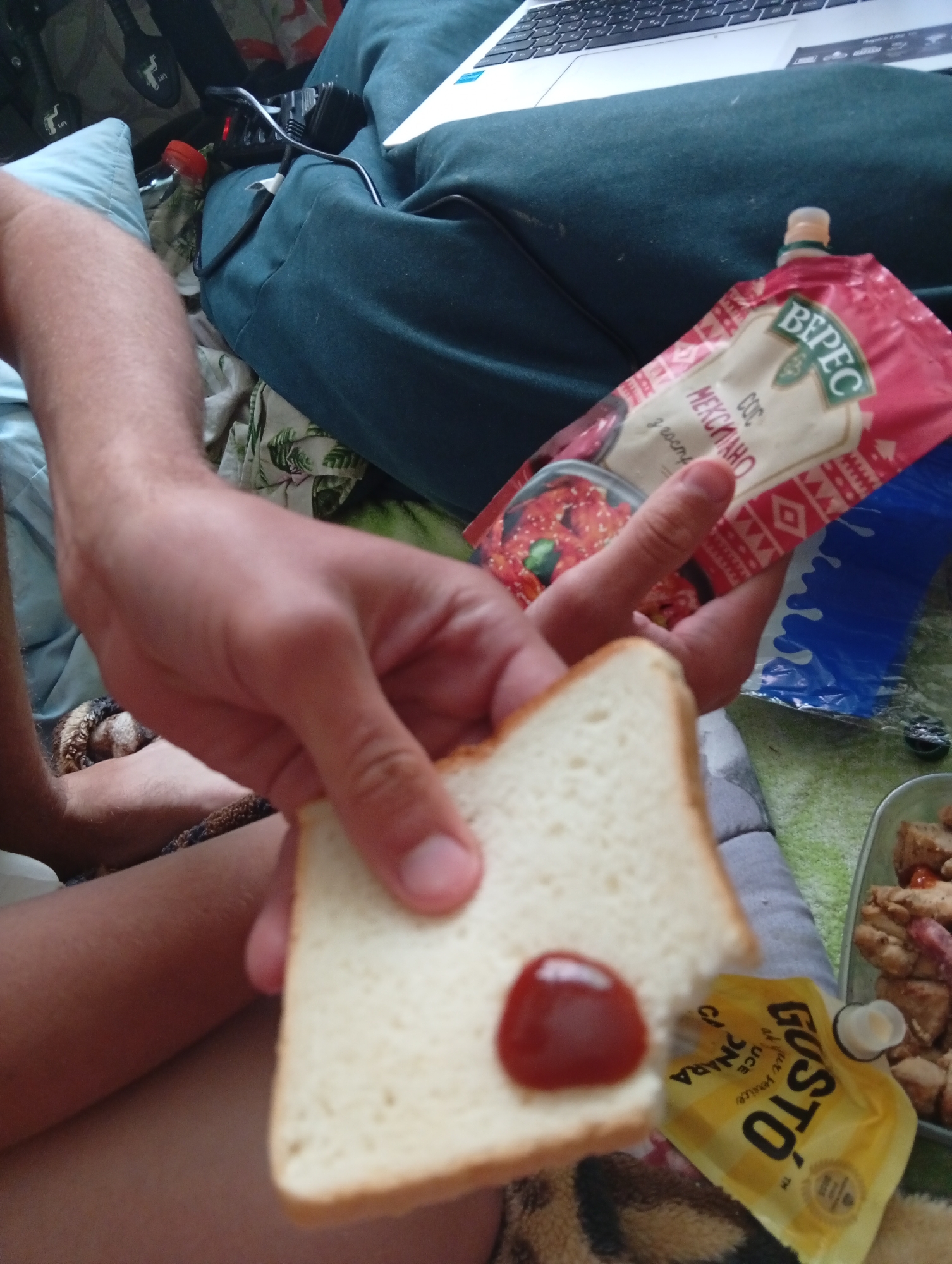 A person is holding a slice of white bread with a dollop of red sauce in their left hand. In their right hand, they are holding a red packet of "Верес" Mexican-style sauce. Adjacent to the bread, there is a yellow packet labeled "GUSTO". Next to it, a metal container appears to have chicken. The person is sitting on a couch in what may be a residential setting. A laptop is partially visible in the top-right corner. The specific location within Poltava, Ukraine, cannot be determined from the image.