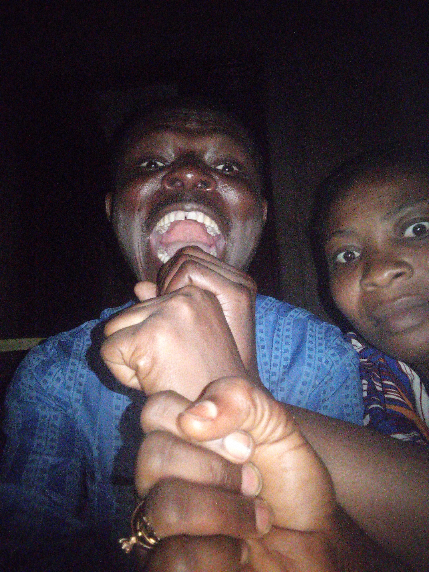 The image shows two individuals in what appears to be a dimly lit indoor setting. The person in the foreground is a male, captured from the chest up, with his mouth wide open in a yell or laugh. He is wearing a blue patterned shirt. His hands are raised, with one hand near his mouth and the other in front of him, partially obscuring his face. A gold ring is visible on one of his fingers. To the right and slightly behind him is another individual, a female, whose face is also visible. She is looking directly at the camera. The background is dark, suggesting a low-light environment. The overall impression is that of a candid, spontaneous moment. Without further information, the specific location in Tempure, Nigeria, cannot be determined.
