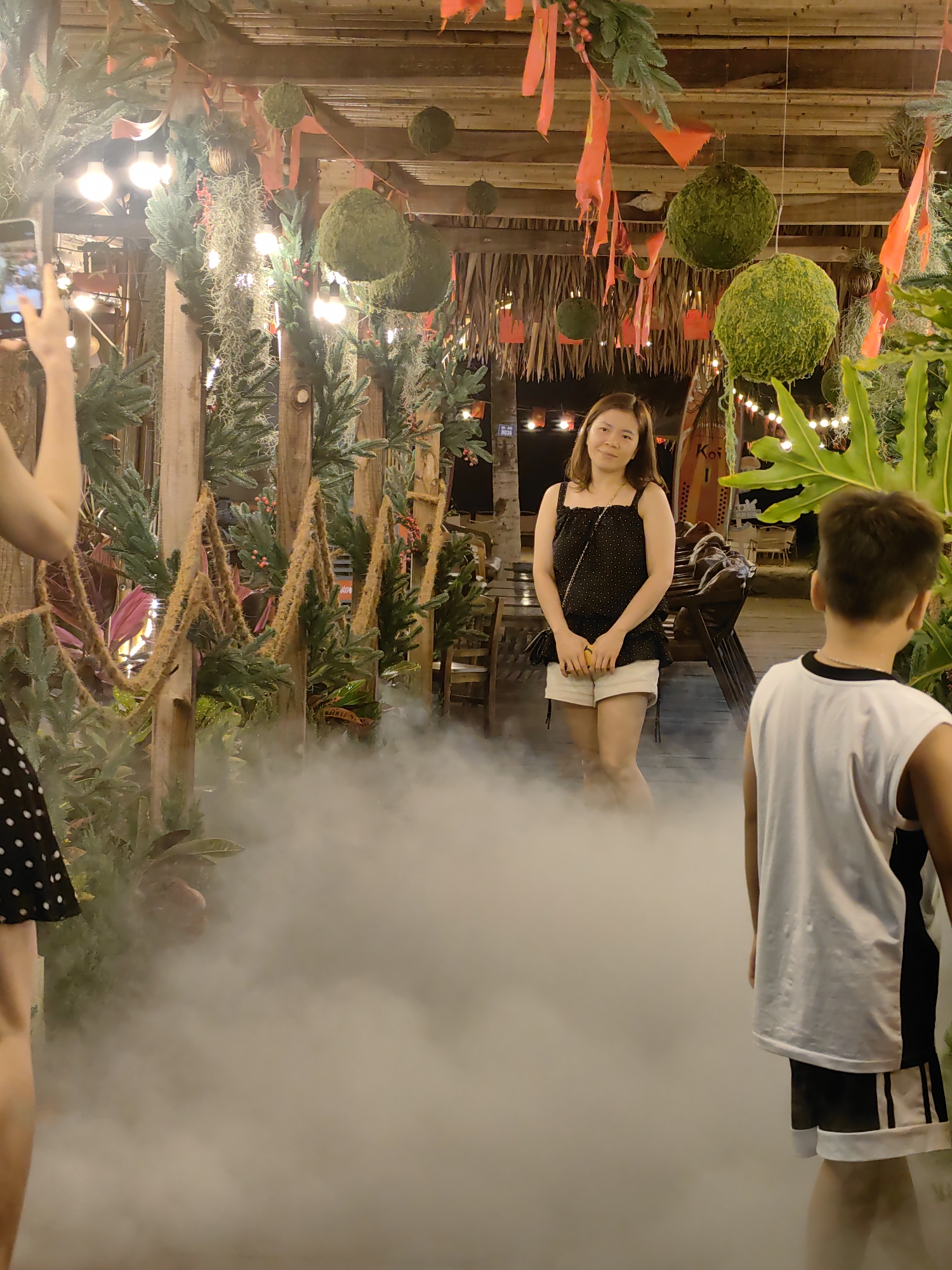Scene in Bai Chay Ward, Vietnam, possibly outdoors, at night. A woman in a black polka-dot top and white shorts stands in the middle ground, smiling toward the camera. To her right, a child in a white tank top and black shorts faces away from the camera. To her left, a person is partially visible, holding a phone up. The background contains foliage, hanging decorative balls, and string lights, and a thatched roof overhead. A layer of fog covers the ground.