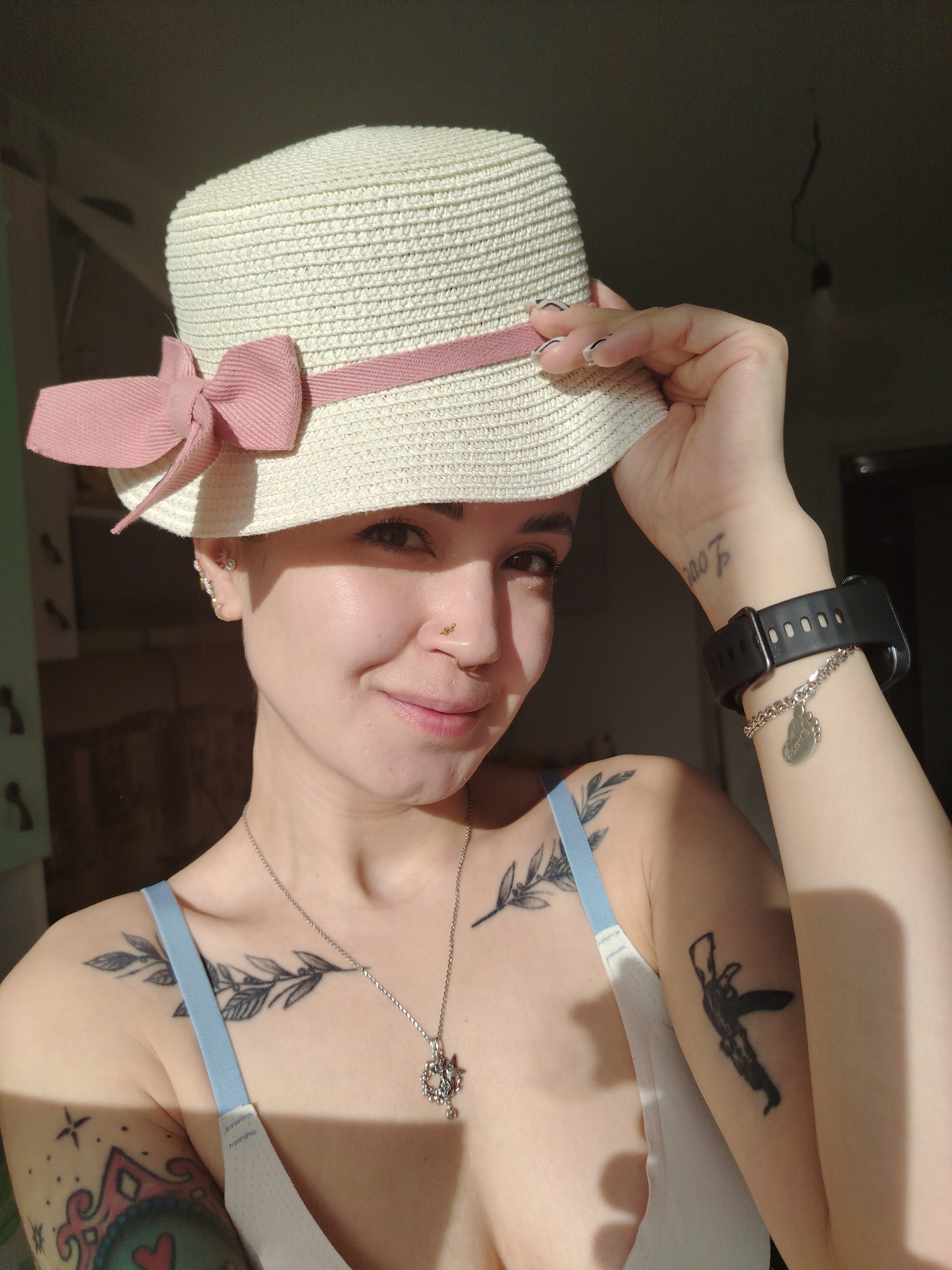A young woman with short hair and a gentle smile is captured in a close-up indoor portrait, likely in Noviy, Russia. She wears a light-colored straw hat adorned with a pink ribbon and bow, which she holds with her left hand. Her face is illuminated by strong natural light from the left, casting distinct shadows on her right side and the background. She is wearing a light-colored bra or bralette, with a light blue strap visible on her left shoulder.
Notable details include multiple ear piercings and two nose piercings on her right nostril. She sports several tattoos: a leafy design on her left chest/shoulder, a colorful, abstract tattoo with a heart on her inner left bicep, and a dark, silhouette-style image on her right upper arm. On her left wrist, she has a small text tattoo resembling "Love" and wears a black smartwatch. A silver-colored chain bracelet with a heart-shaped charm, faintly engraved with what appears to be "Mom", is worn below the watch. A silver necklace with a winged pendant is also visible around her neck. The background is dark and out of focus, suggesting a private indoor setting during daytime hours, with the sun creating a strong contrast. She looks directly at the camera, conveying a relaxed and pleasant demeanor.