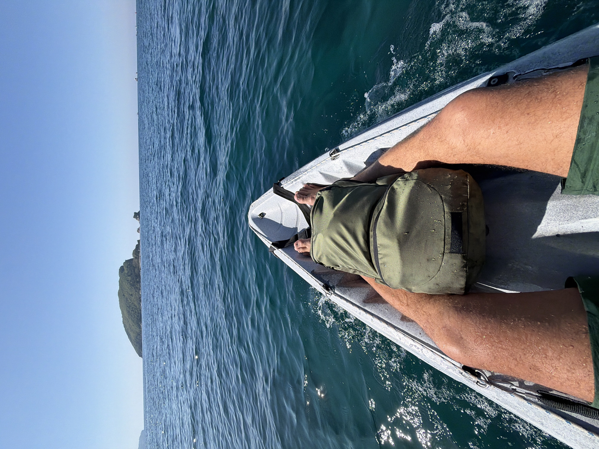 A person is sitting in a gray kayak on the Adriatic Sea. The person has a green bag on their lap. They are wearing green shorts. The water is blue with visible sunlight reflections. A distant land mass is visible in the left side of the image. The image was likely taken in Budva, Montenegro.