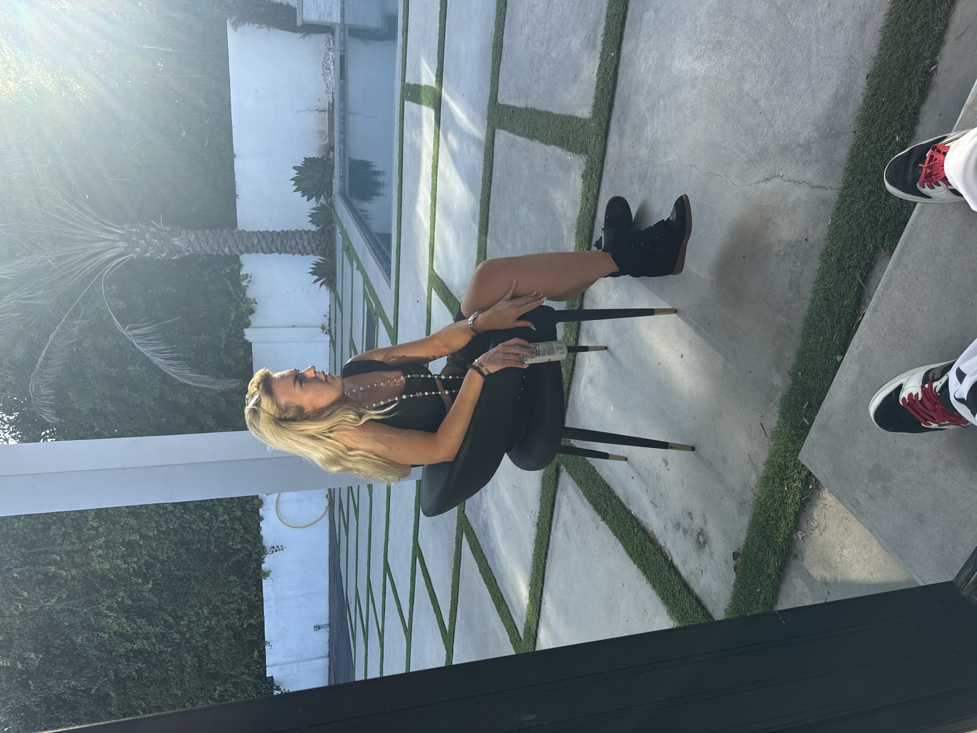 A medium-angle, slightly rotated shot shows a blonde-haired woman in a black outfit seated in a black chair, holding a water bottle in her lap, likely outdoors. The setting seems to be in a backyard, featuring a modern patio design with rectangular concrete slabs separated by strips of grass. A rectangular swimming pool is visible in the background, as well as a palm tree and lush greenery. The strong sunlight suggests the image was taken during the day. Another person's feet wearing sneakers are visible on the right side of the frame. The overall impression is that of an affluent, outdoor location. Due to the types of landscaping and architecture displayed, the setting may be in Los Angeles, United States of America.