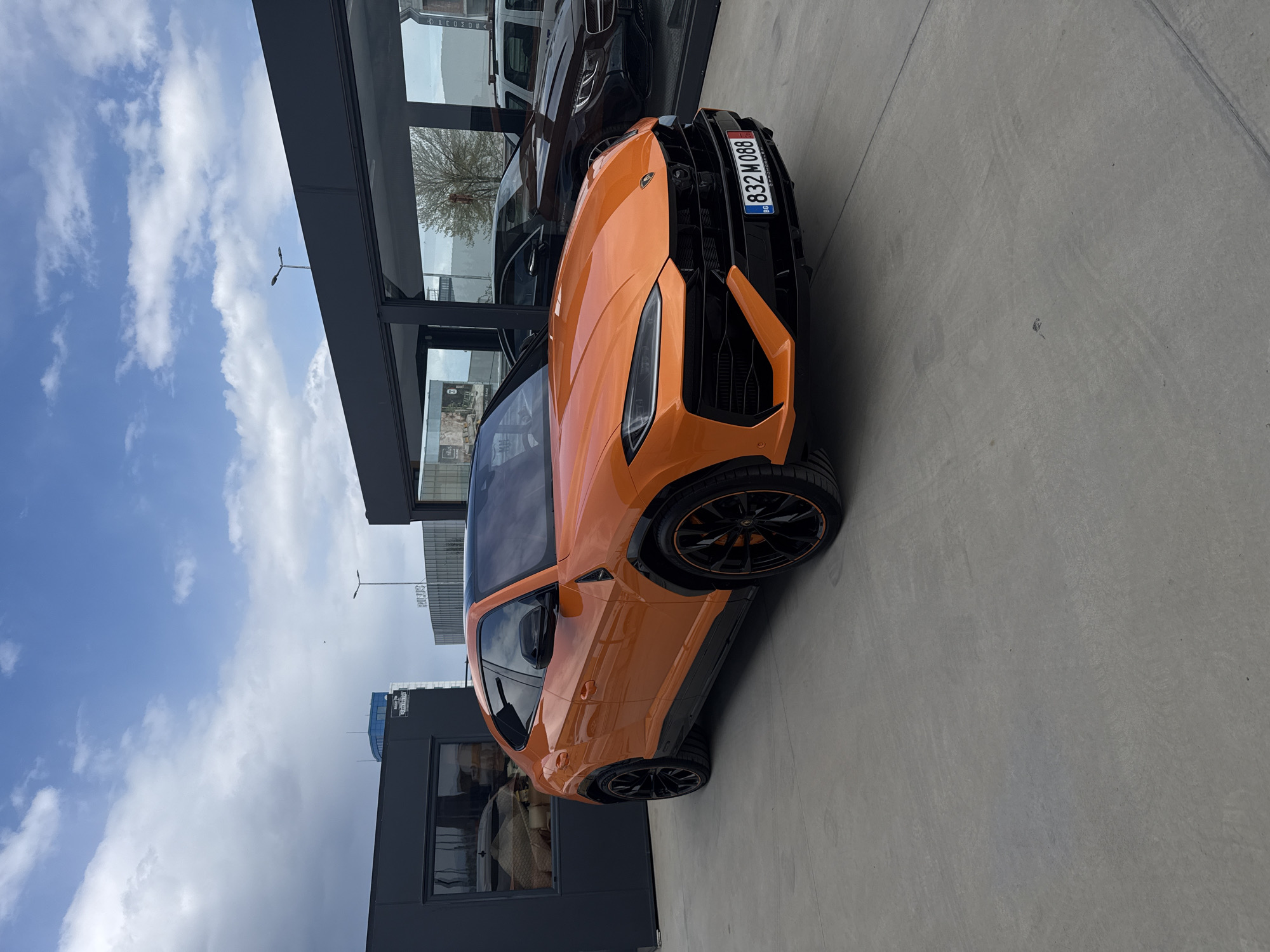 The image shows an orange Lamborghini Urus parked in front of a building in Plovdiv, Bulgaria. The license plate number is 832 M 088. The car is parked on a concrete surface. The building has glass windows and a black frame. There are other cars visible in the background through the windows of the building. The sky is blue with some clouds.