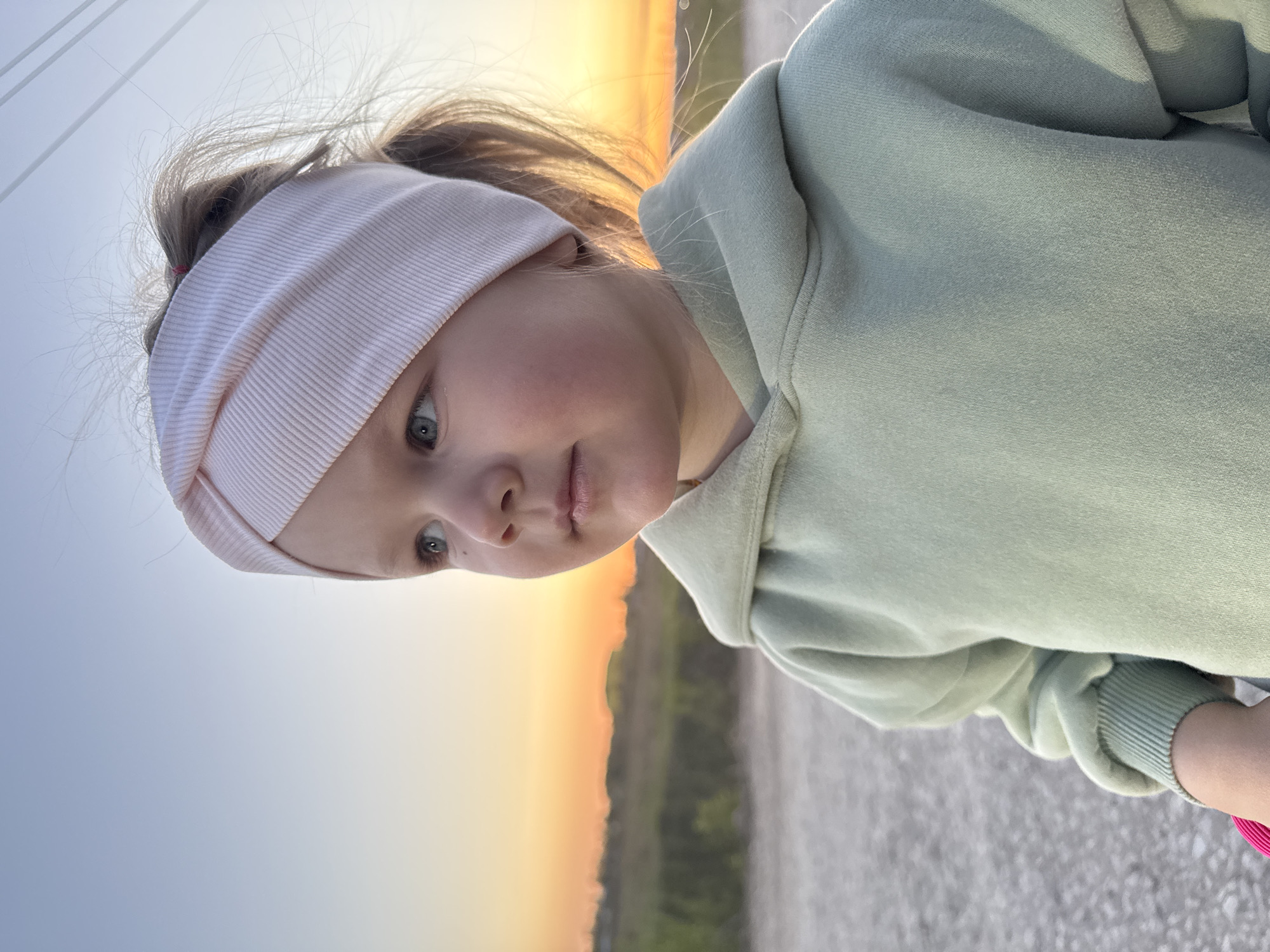 A young girl is standing outdoors during a sunset. She wears a light green hooded sweatshirt and a pink headband. The background includes the fading light of the sunset, a field, and what appears to be a road or path. The location cannot be conclusively determined as Горный, Russia, as there are no specific identifying features in the photo to confirm this.