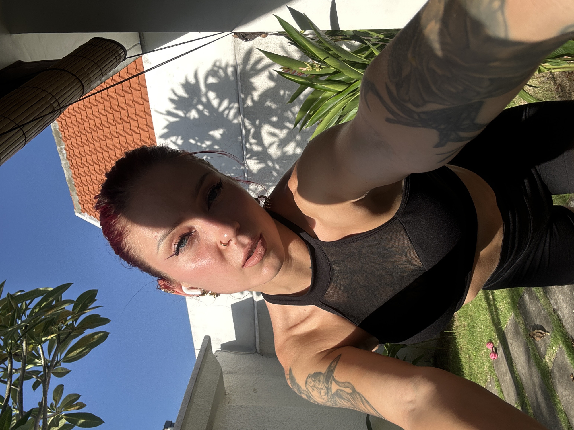 A medium shot captures a woman facing the camera, presumably taking a selfie. She has fair skin, red hair pulled back, and distinct eye makeup. She is wearing a black sports bra with mesh detailing at the top and black leggings. Her left arm, visible to the right, is adorned with a tattoo.
The setting is outdoors, with elements indicating a garden or yard area. Background details include green plants and a tree with green leaves on the left side. Architectural features such as a roof with orange tiles and a white wall are also visible. The shadows suggest it's a sunny day.
Based on information from the metadata, the location is North Kuta, Bali, Indonesia.